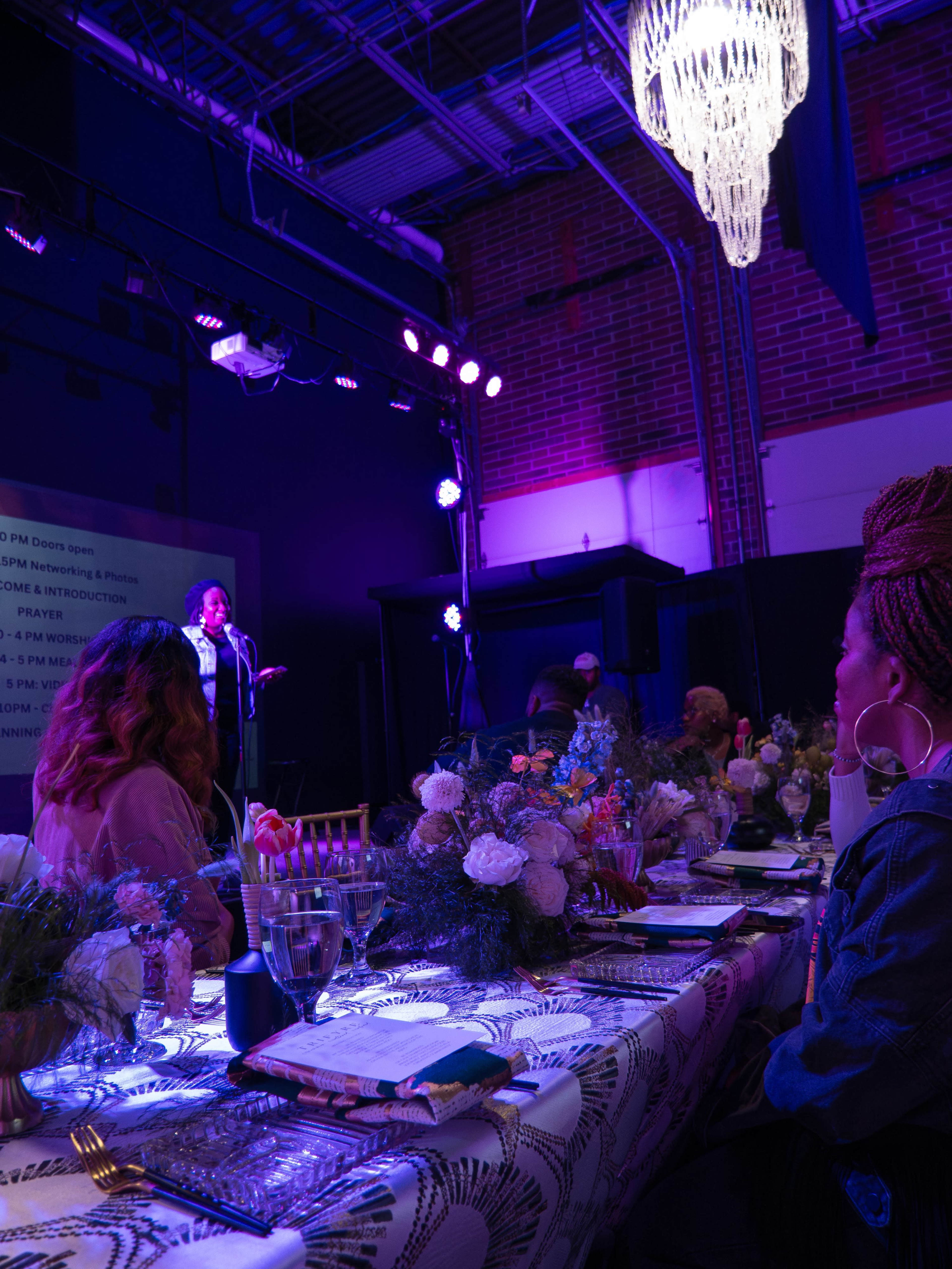 A performer stands on stage in a dimly lit venue while an audience gathers around a long table decorated with flowers and set for a meal.