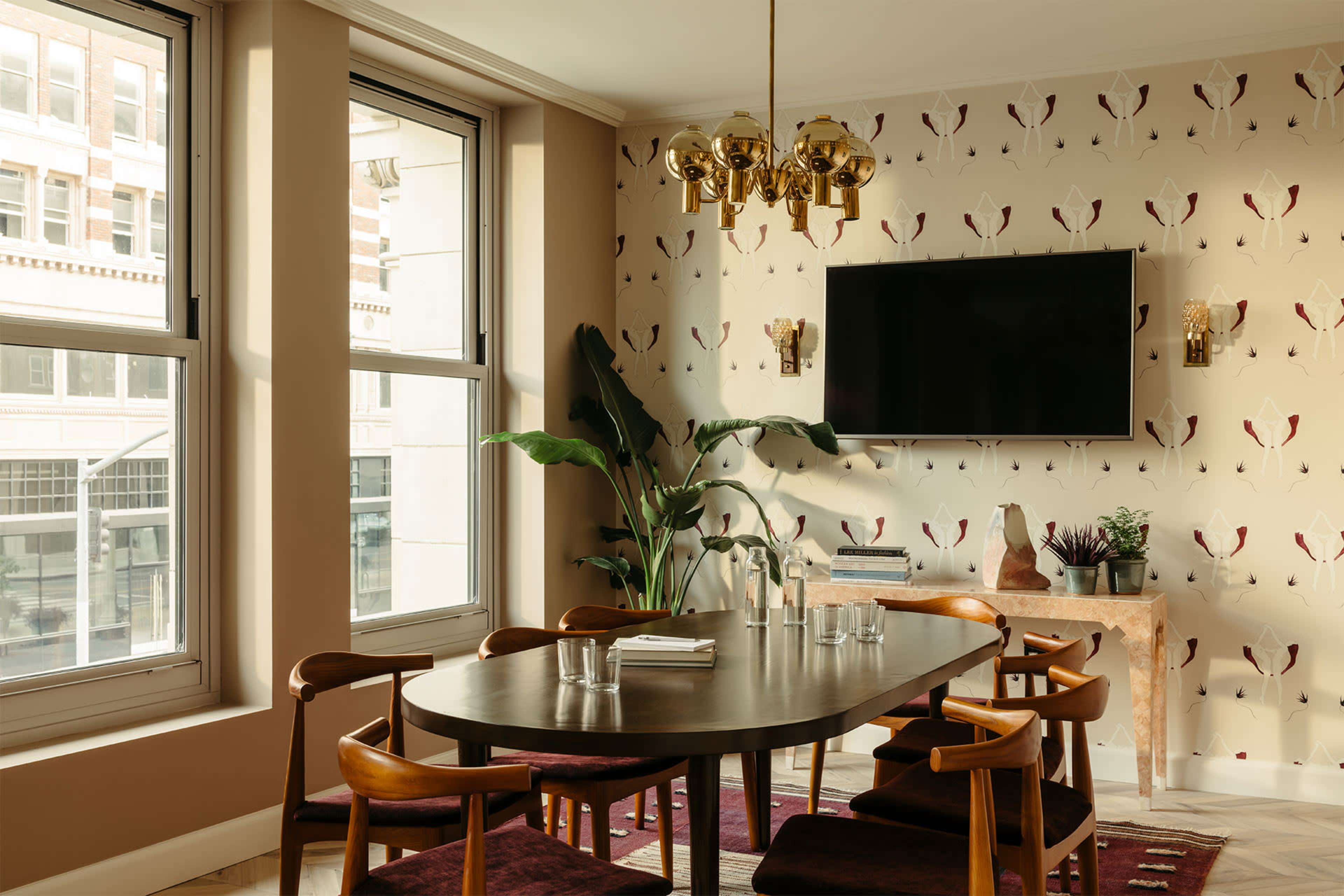 A dining room features a round table surrounded by chairs, against a backdrop of patterned wallpaper and a mounted television.