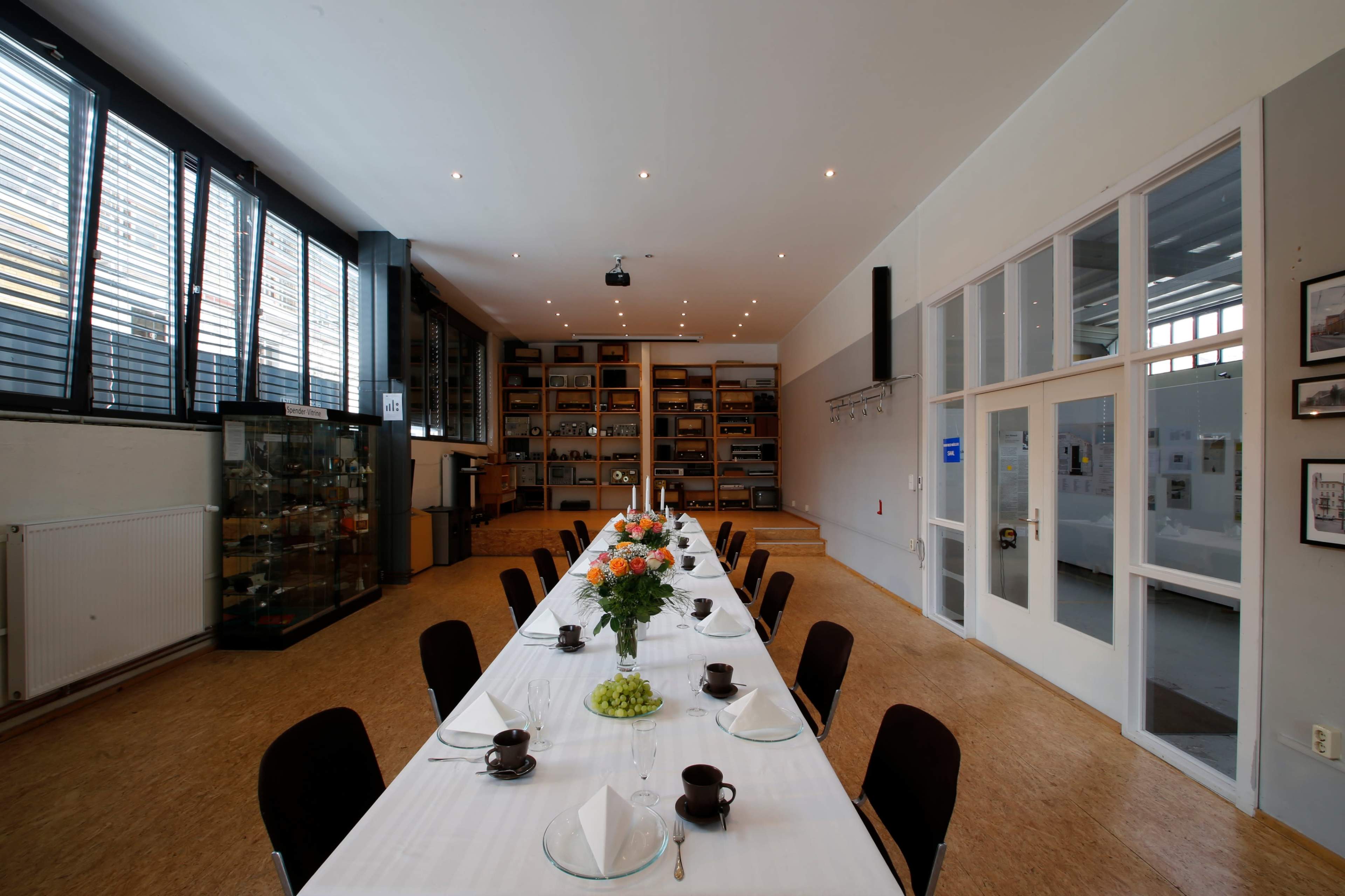 A long dining table is set with floral arrangements and tableware in a well-lit, modern room featuring shelves and display cases.