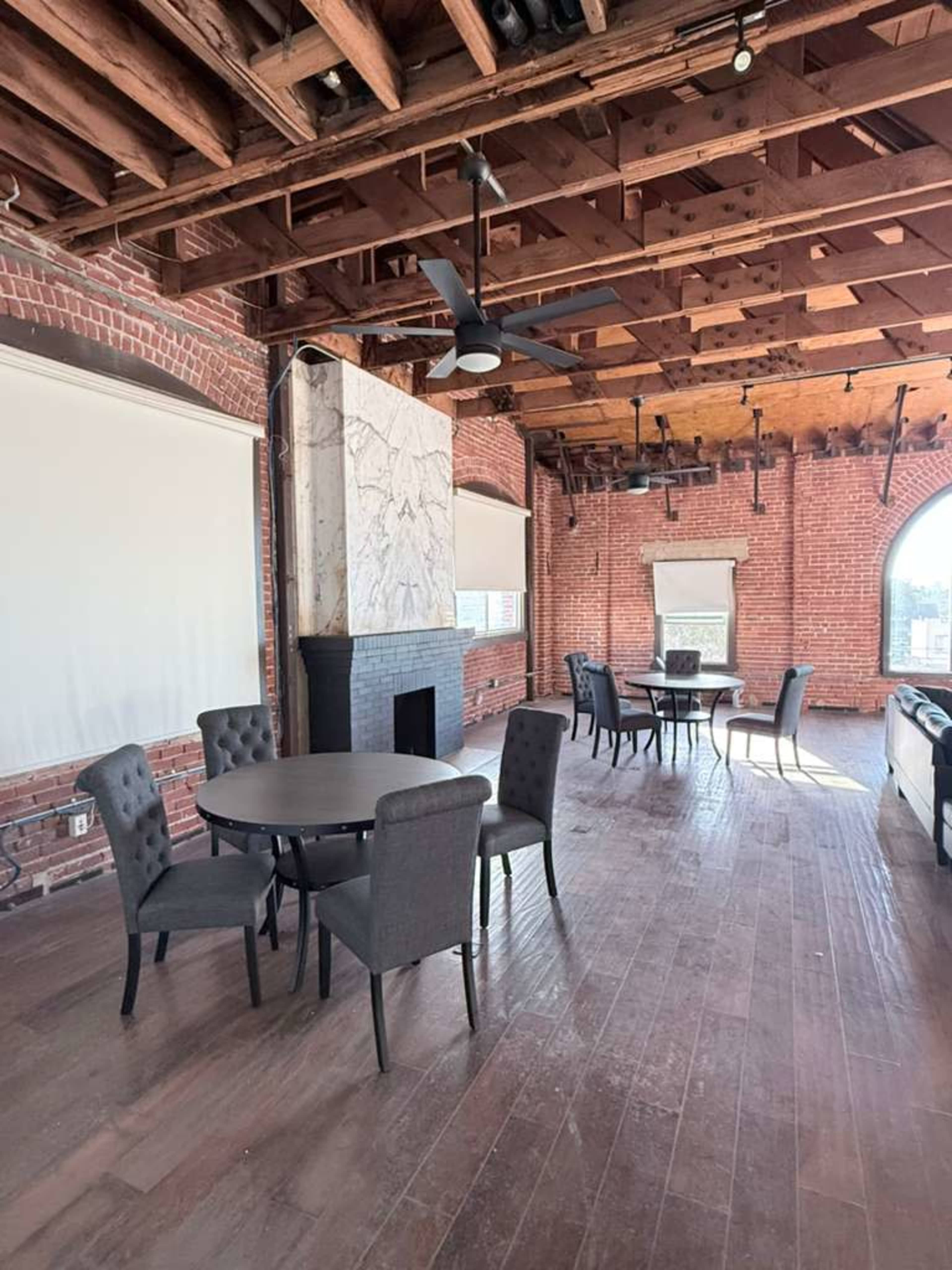 The image shows an open interior space with exposed wooden beams, brick walls, and several black chairs arranged around round tables.