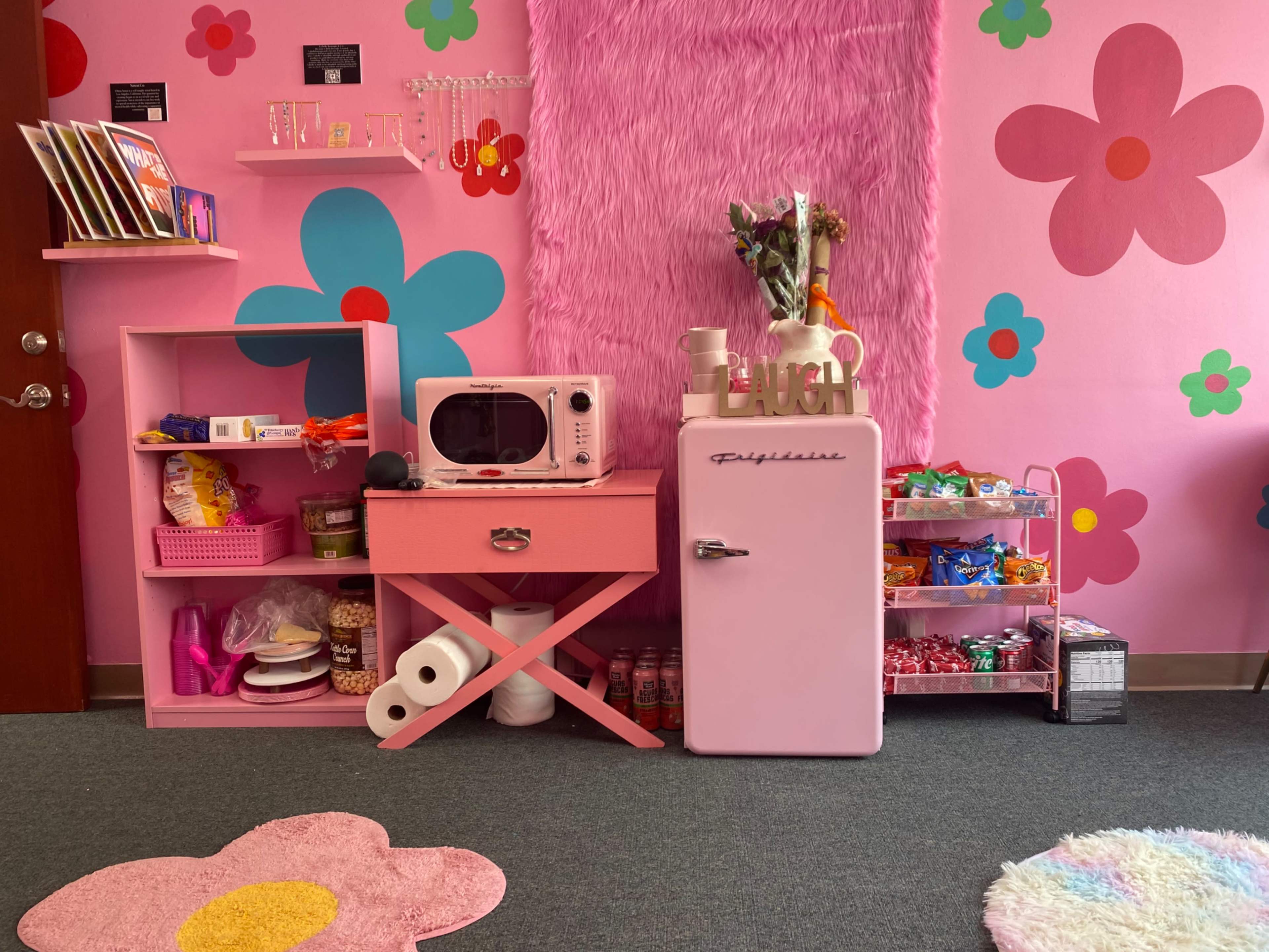 The image shows a colorful room with pink walls decorated with flowers, featuring a pink refrigerator, a pink microwave on a table, and various snacks arranged on shelves.