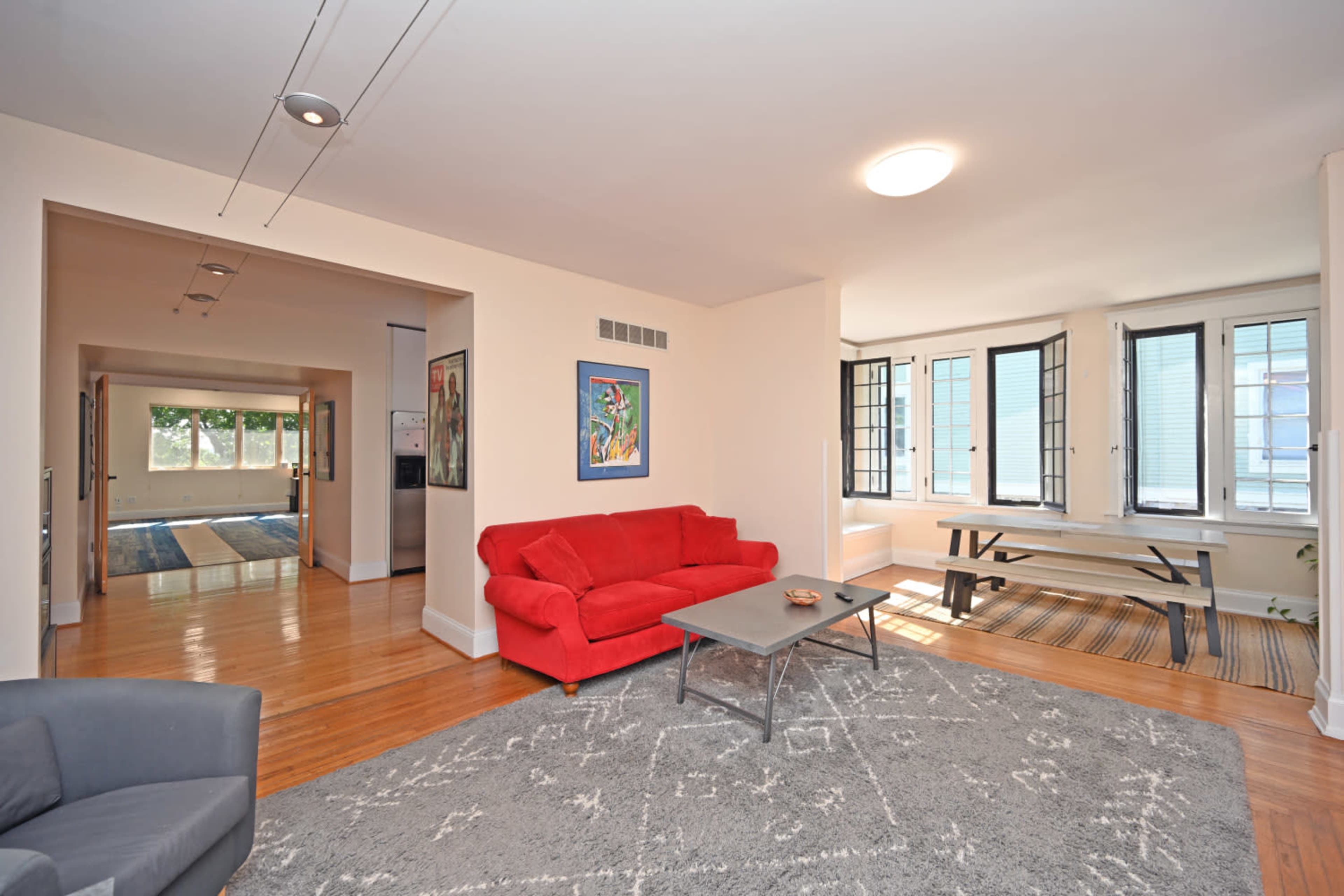 The image shows a spacious living area featuring a red sofa, a gray rug, a coffee table, and large windows allowing natural light.