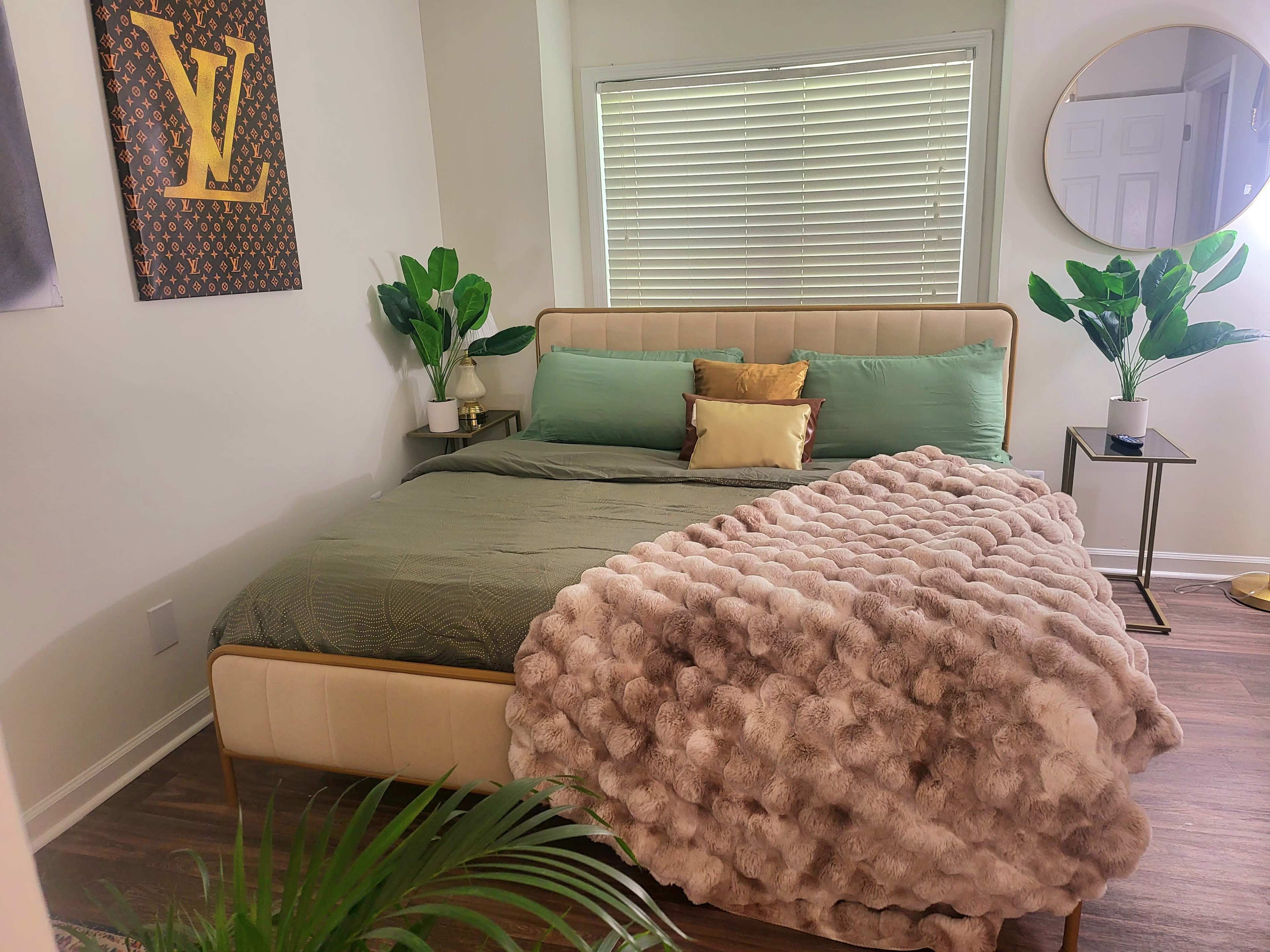 A neatly made bed with green pillows and a plush, textured blanket in a well-lit bedroom featuring plants and a decorative wall art.