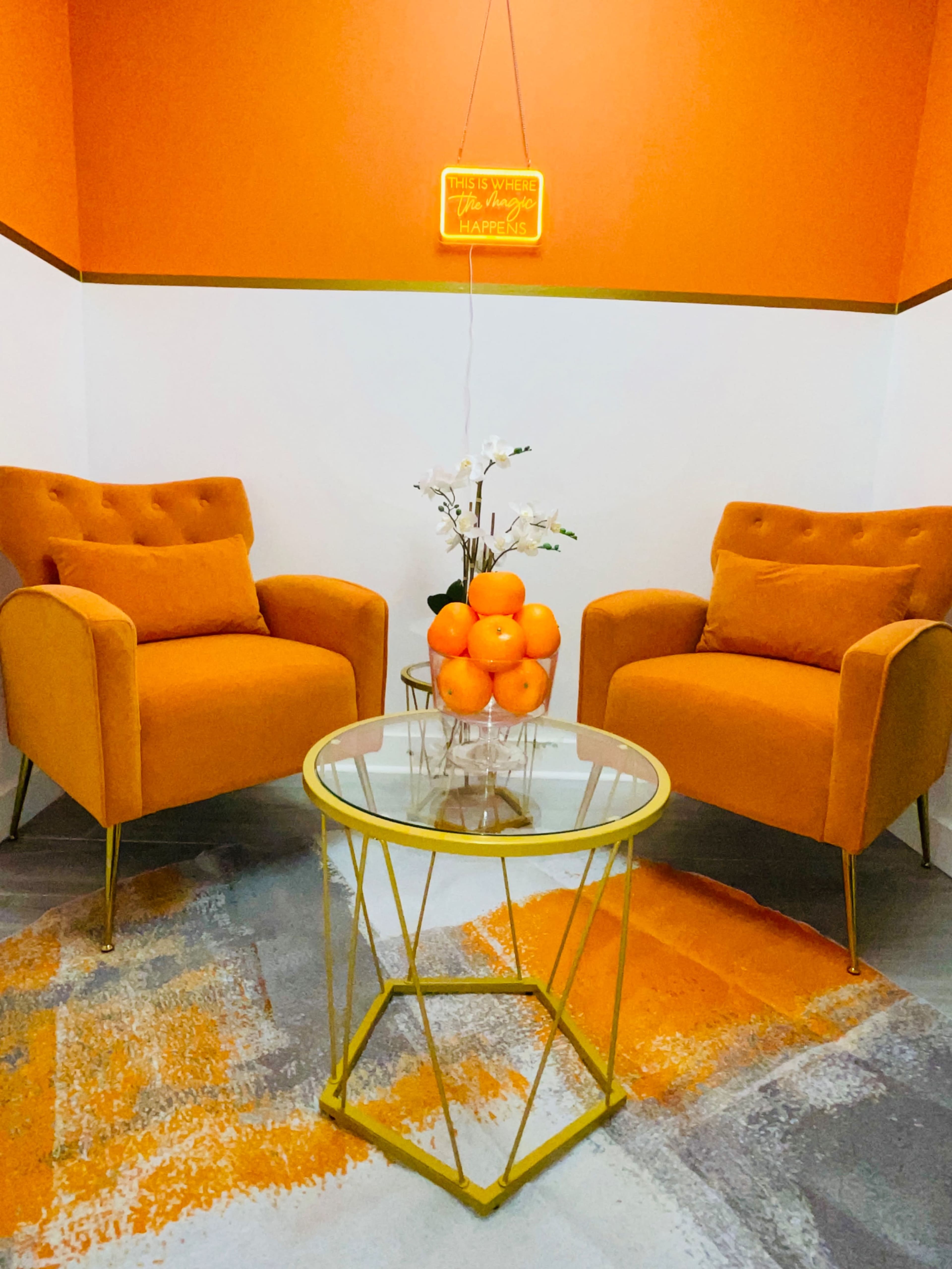The scene features two orange armchairs facing a glass table with a bowl of oranges on it, all set against an orange and white wall.