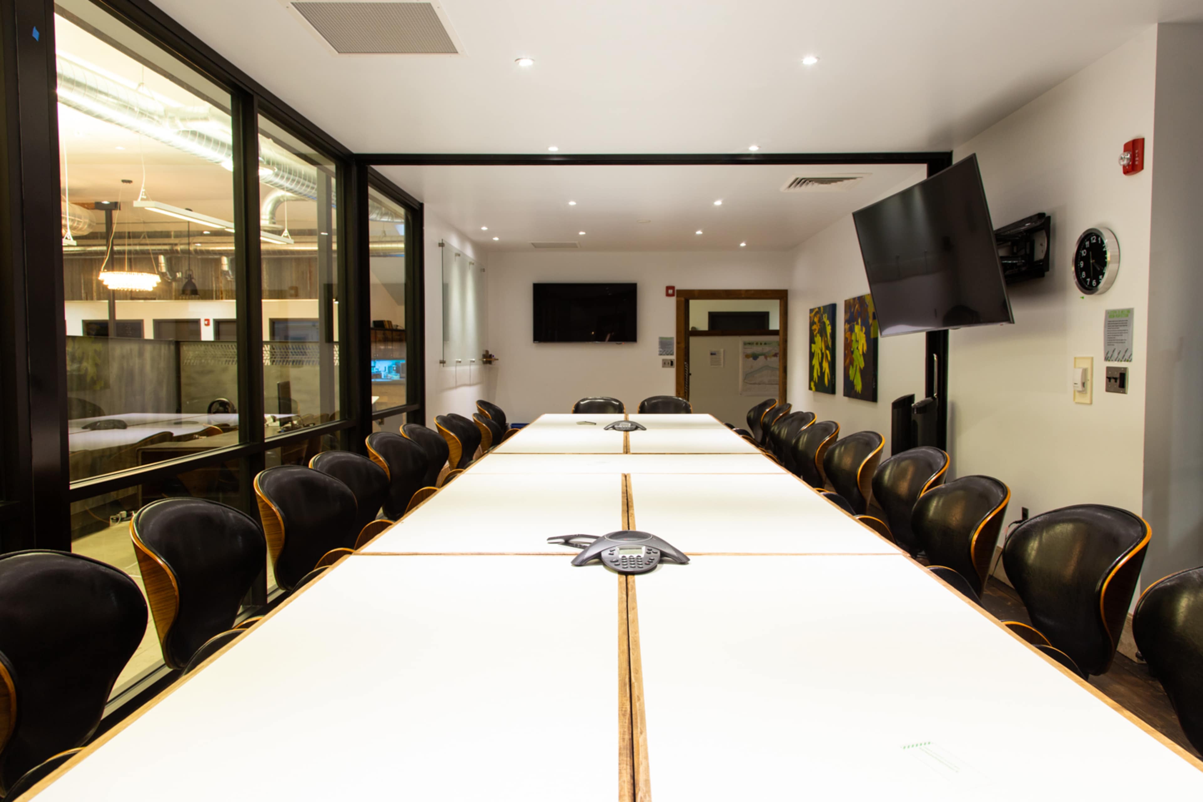 A large conference room features a long table with multiple chairs arranged around it, equipped with a speakerphone and wall-mounted displays.