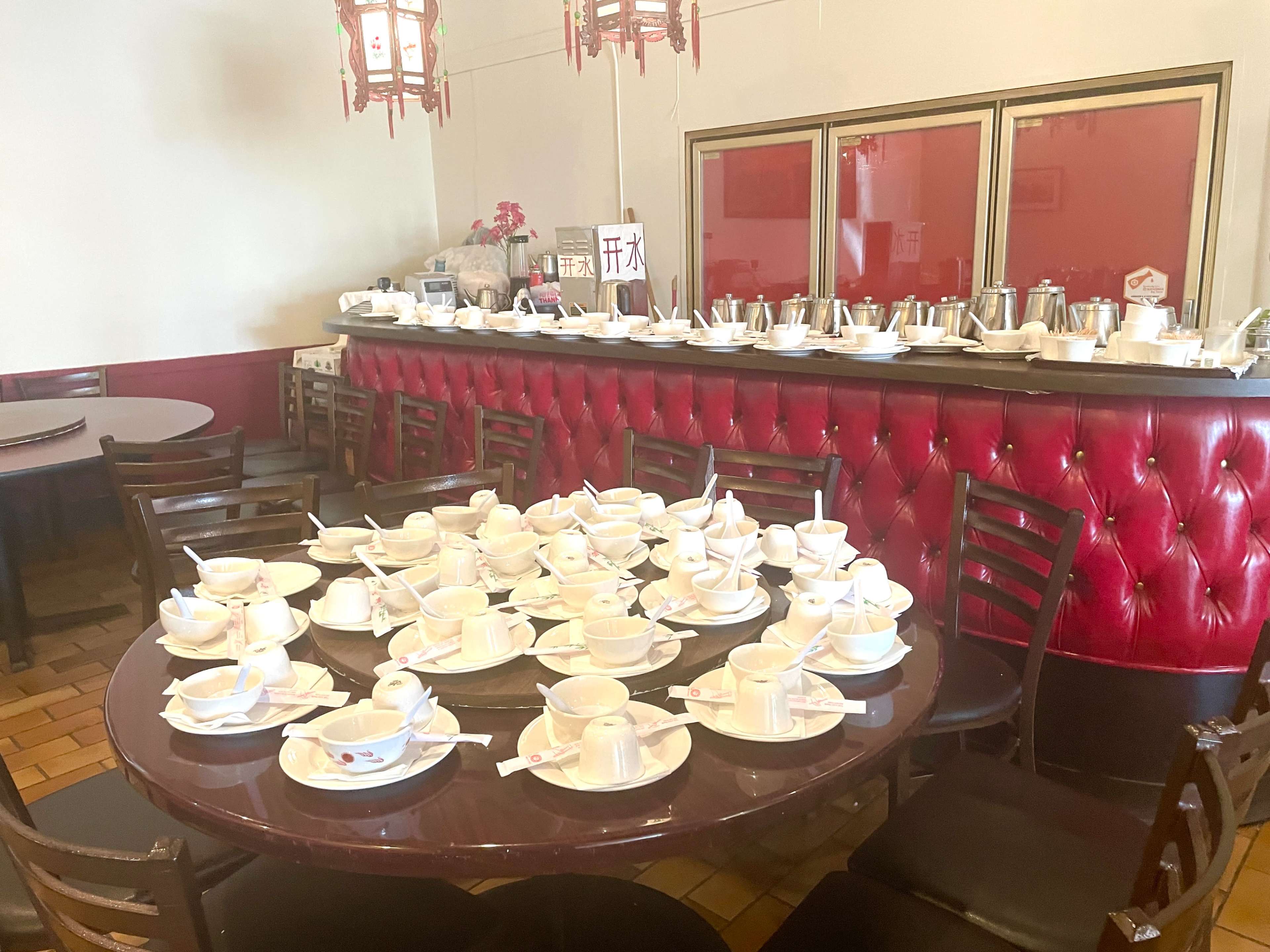A dining area features multiple tables with stacked cups and saucers, while a red leather bench lines one wall.