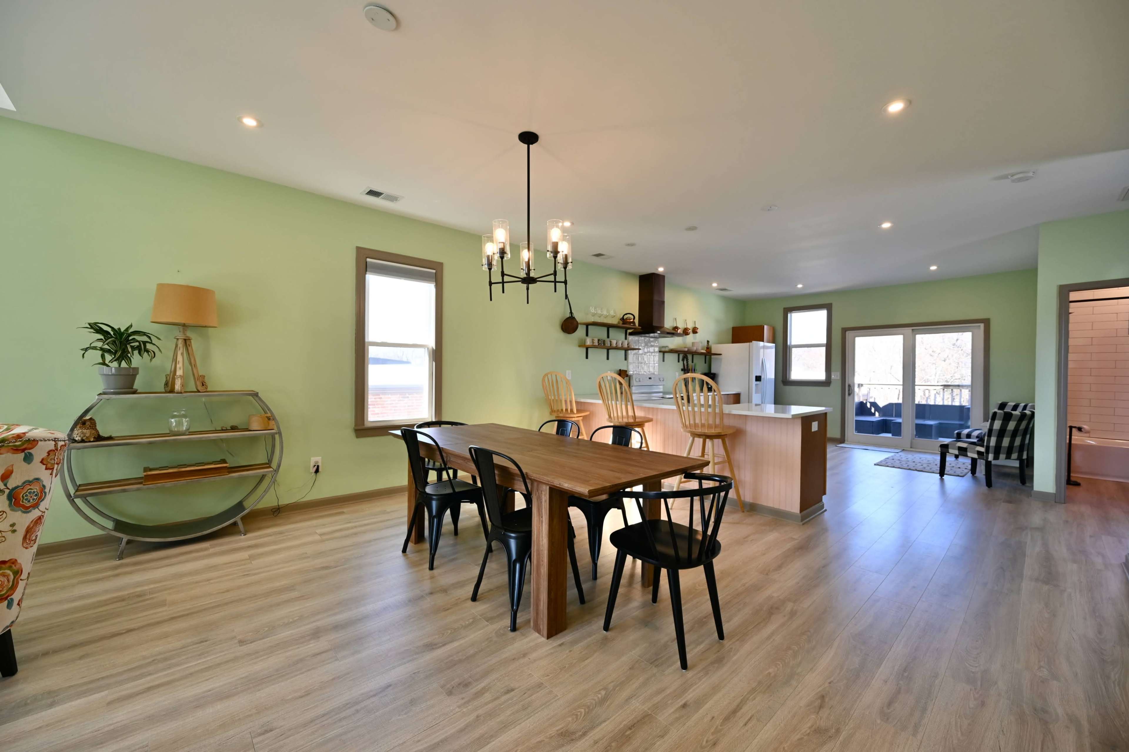 A dining area features a wooden table with black chairs, surrounded by light green walls, and opens to a modern kitchen with a white countertop and appliances.