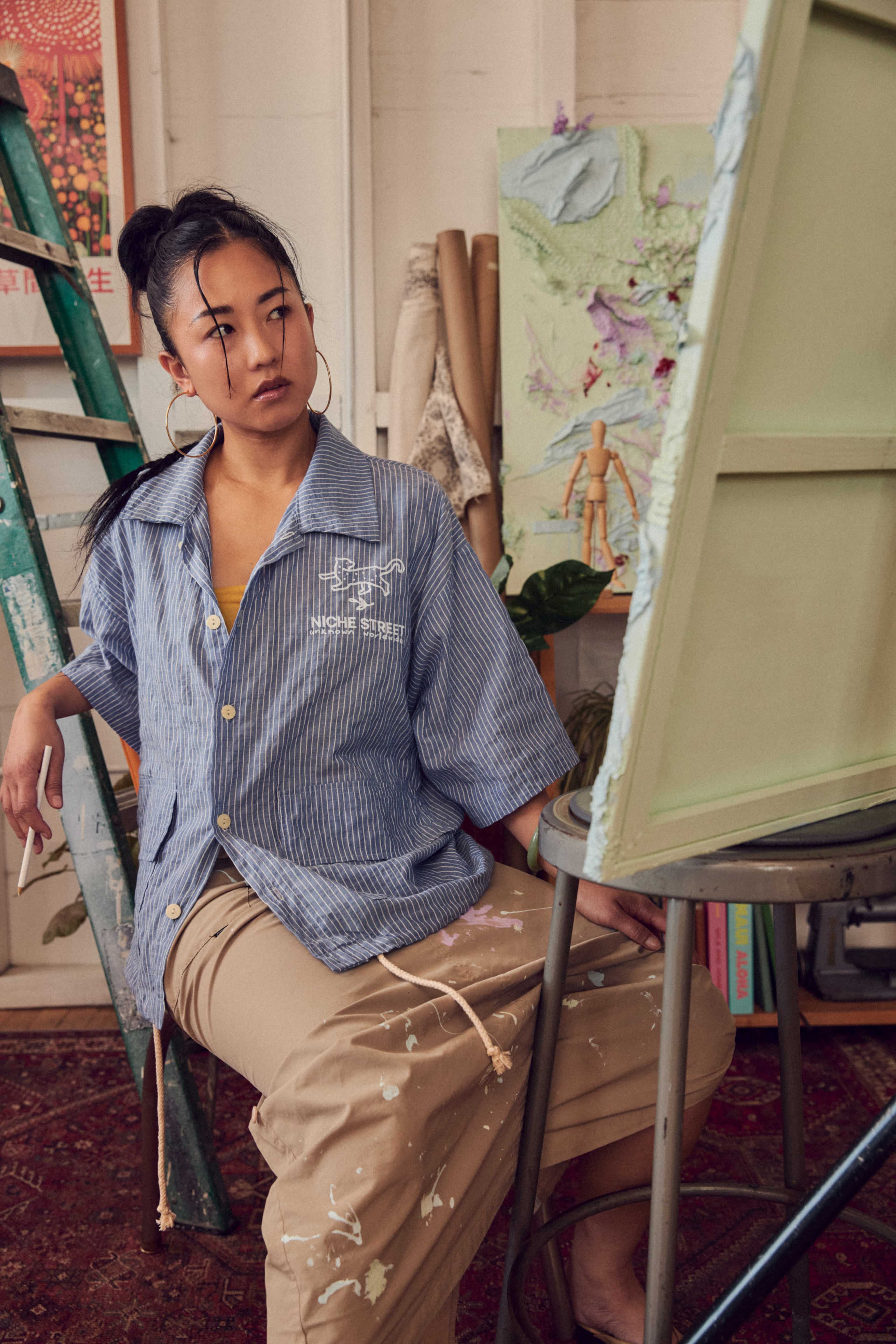 A woman sits on a stool in an artist's studio, wearing a striped shirt and beige pants splattered with paint, while looking thoughtfully toward an easel.