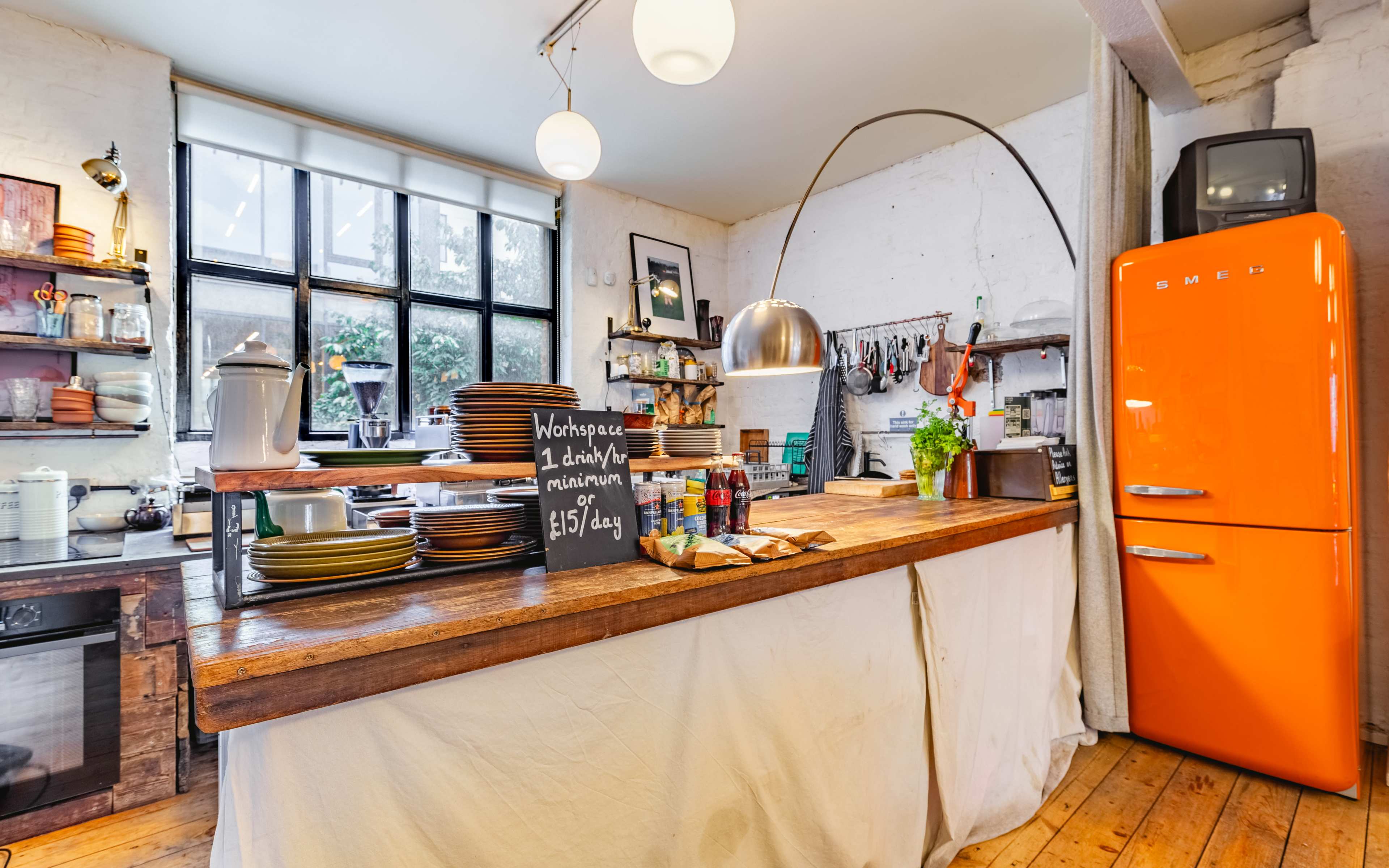 A contemporary kitchen with a wooden island, shelves holding dishware, and an orange retro refrigerator against a white wall.