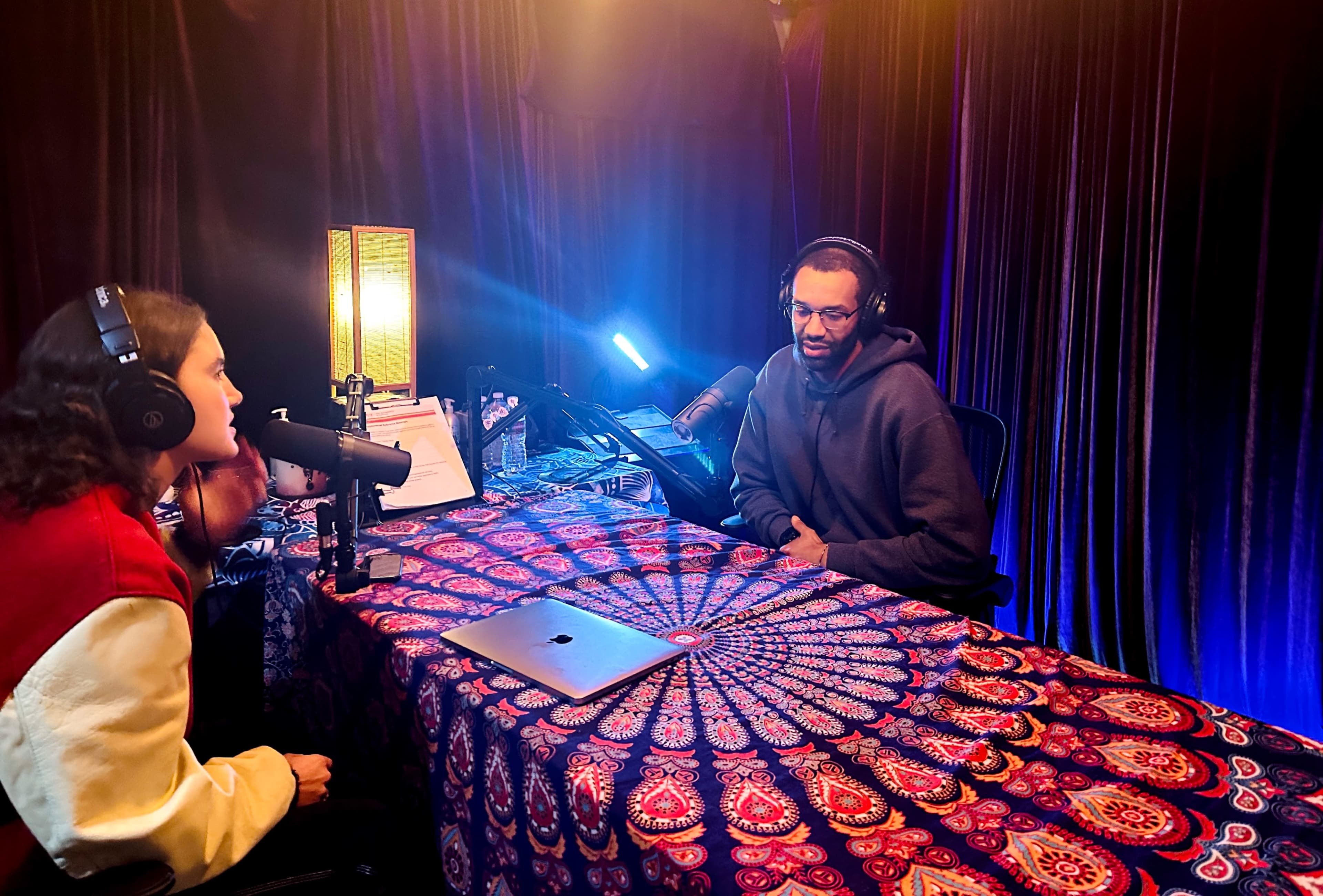 A woman and a man sit at a table covered with a patterned cloth, each wearing headphones and engaged in a conversation in a dimly lit studio.