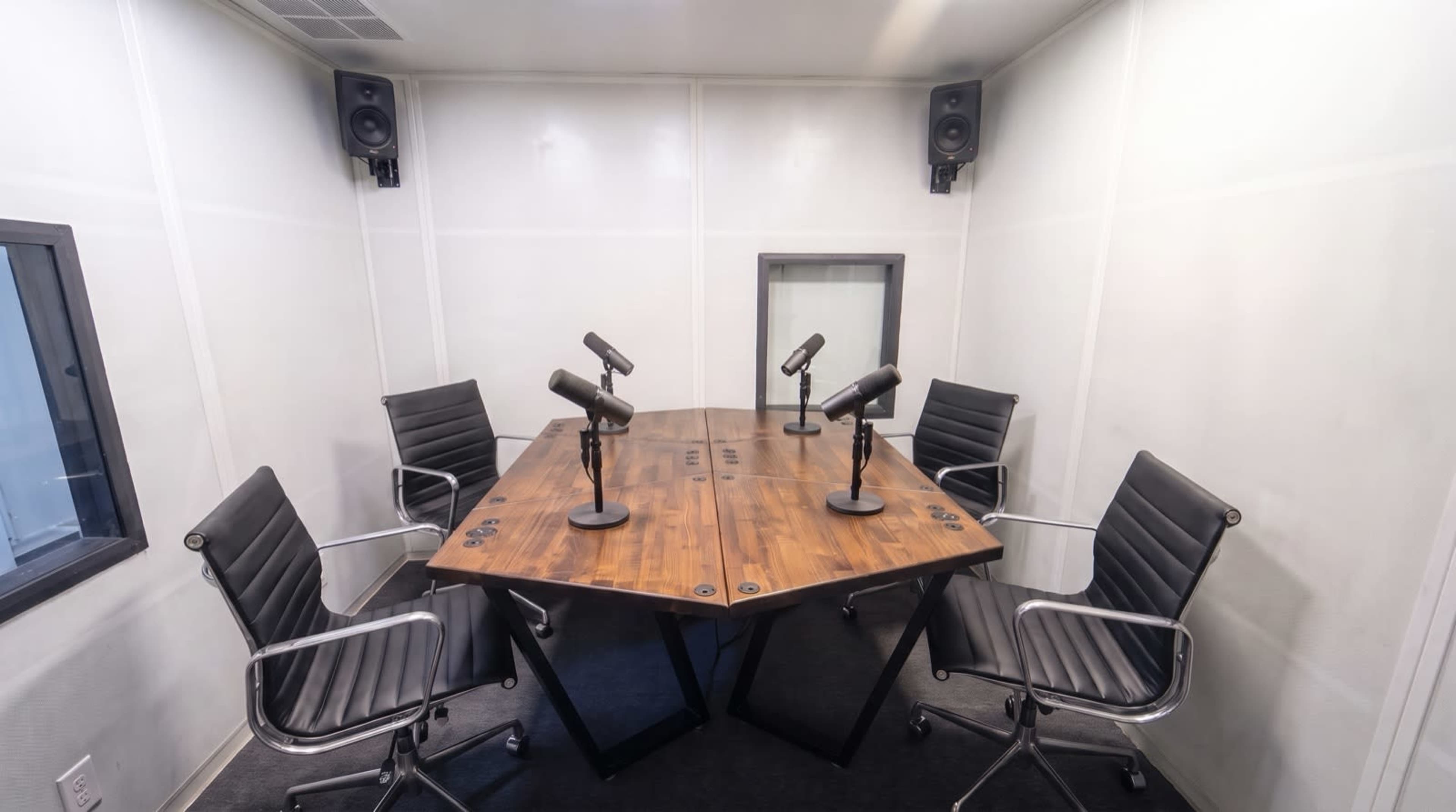 The image shows a soundproof recording studio with a large wooden table and four black ergonomic chairs, each equipped with a microphone.
