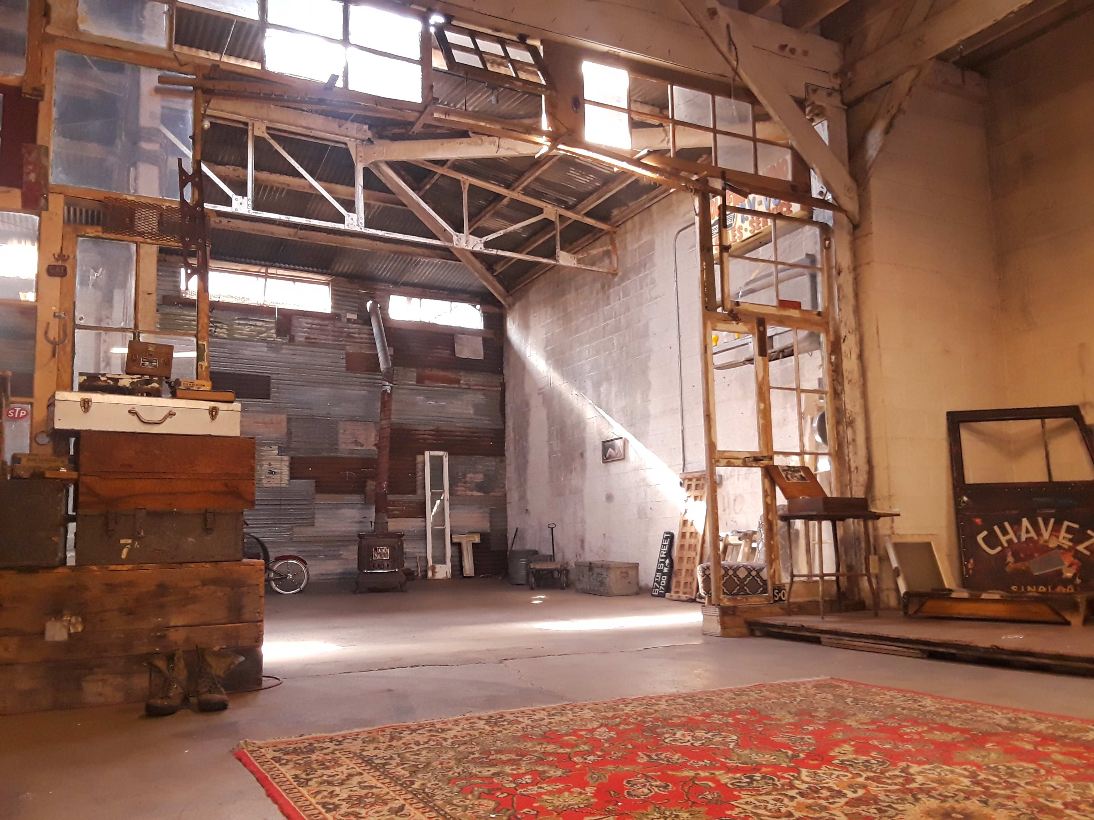 An industrial space with exposed beams, wooden crates, and a patterned rug on the floor, illuminated by natural light coming through large windows.
