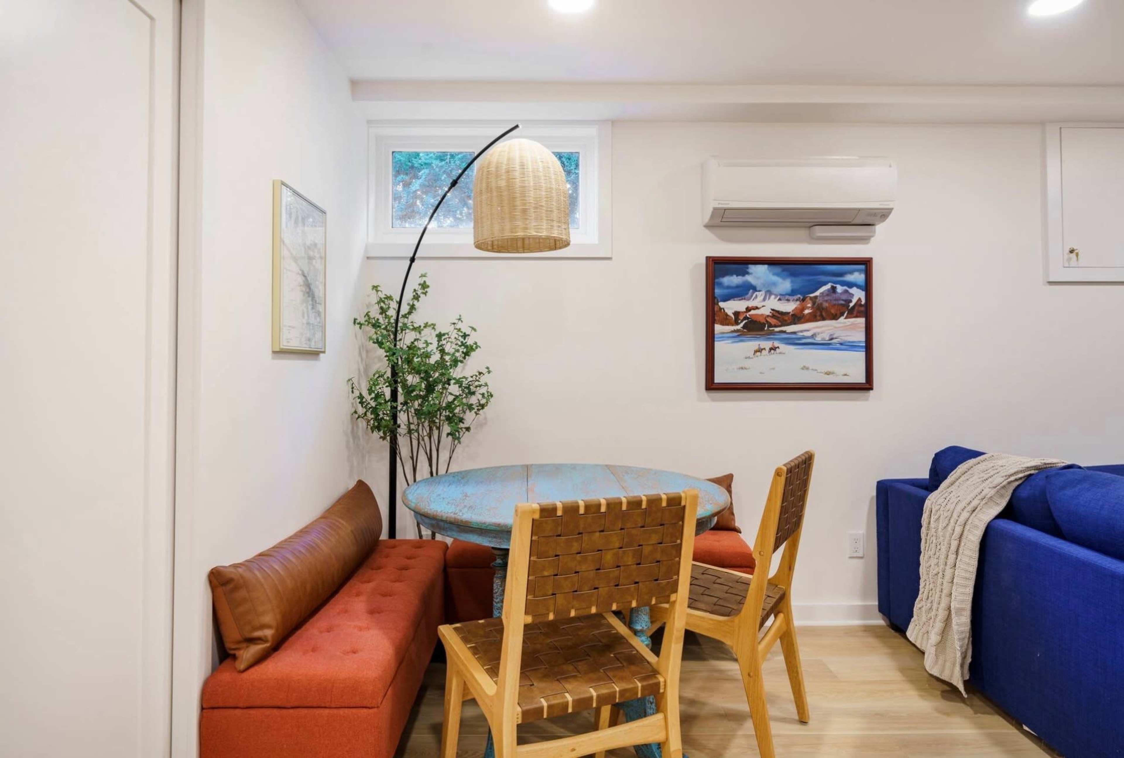 A round wooden table is surrounded by simple wooden chairs and a bench in a bright, minimalist corner of a room with a painting on the wall.