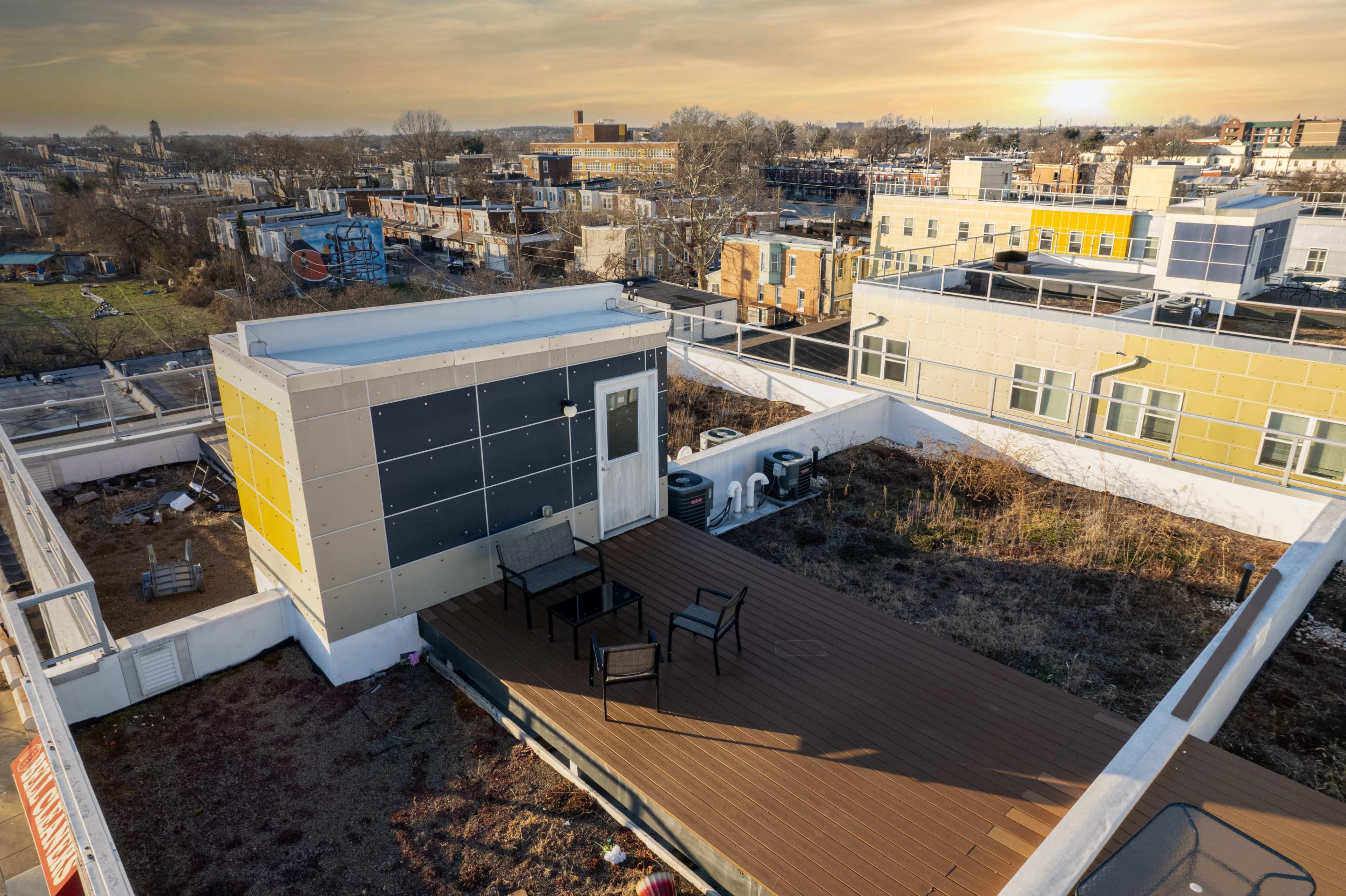 A rooftop deck with seating, surrounded by urban buildings and greenery in the background under a sunset sky.