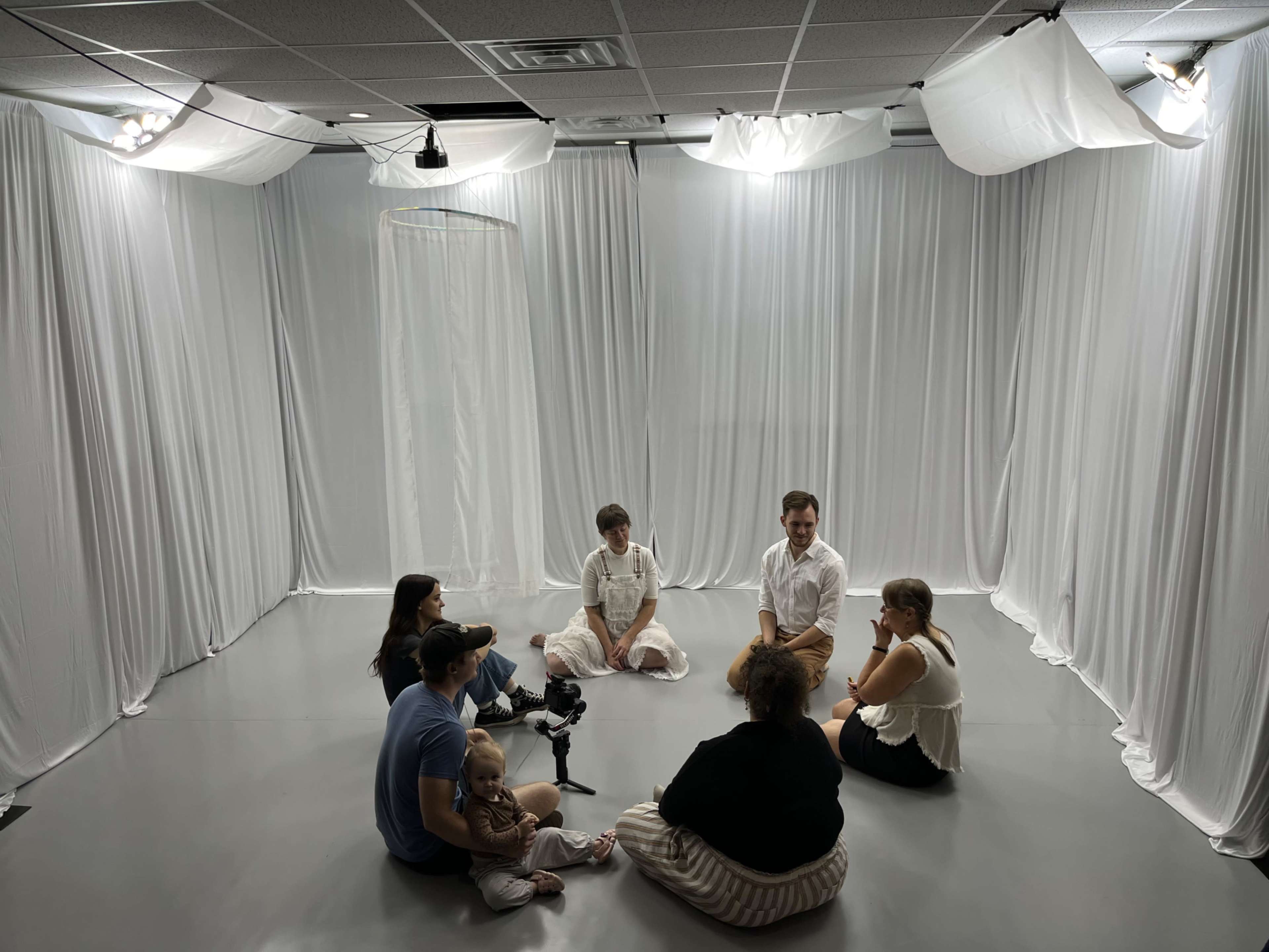 A group of seven people sits in a circle on the floor of a softly lit room with white fabric draping the walls.