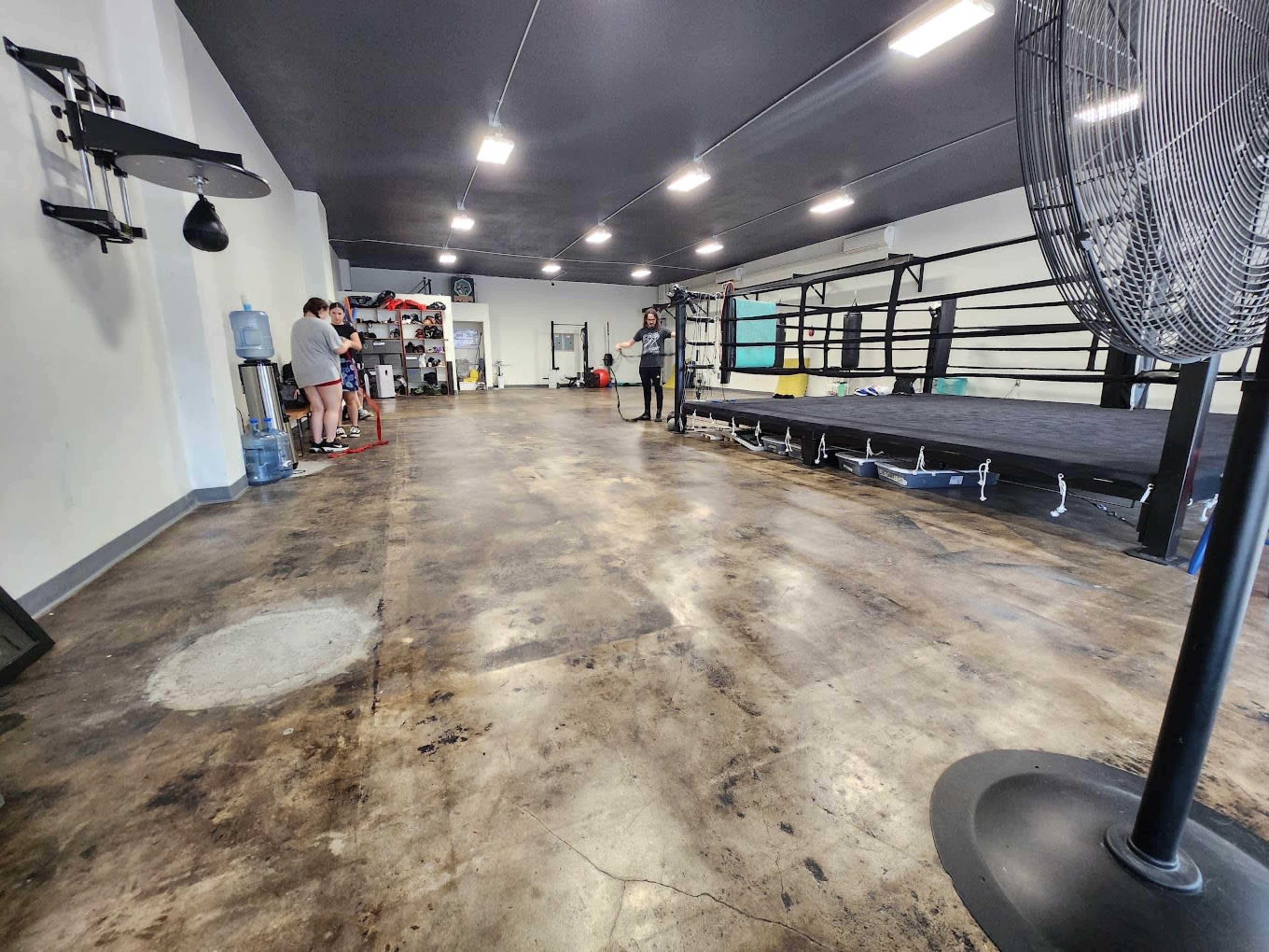 A spacious indoor boxing gym with a ring in the center, workout equipment along the walls, and a few people standing near a water cooler.
