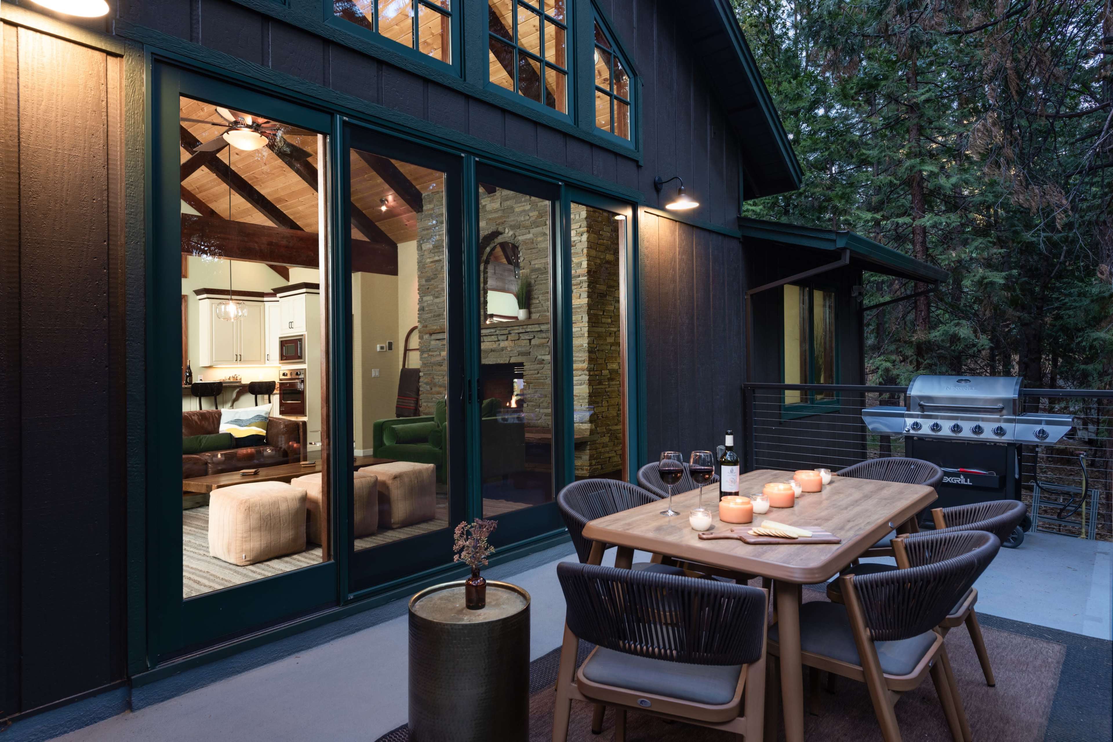 A modern outdoor dining area features a wooden table with candles, surrounded by chairs, adjacent to a house showcasing large windows that reveal a cozy interior space.