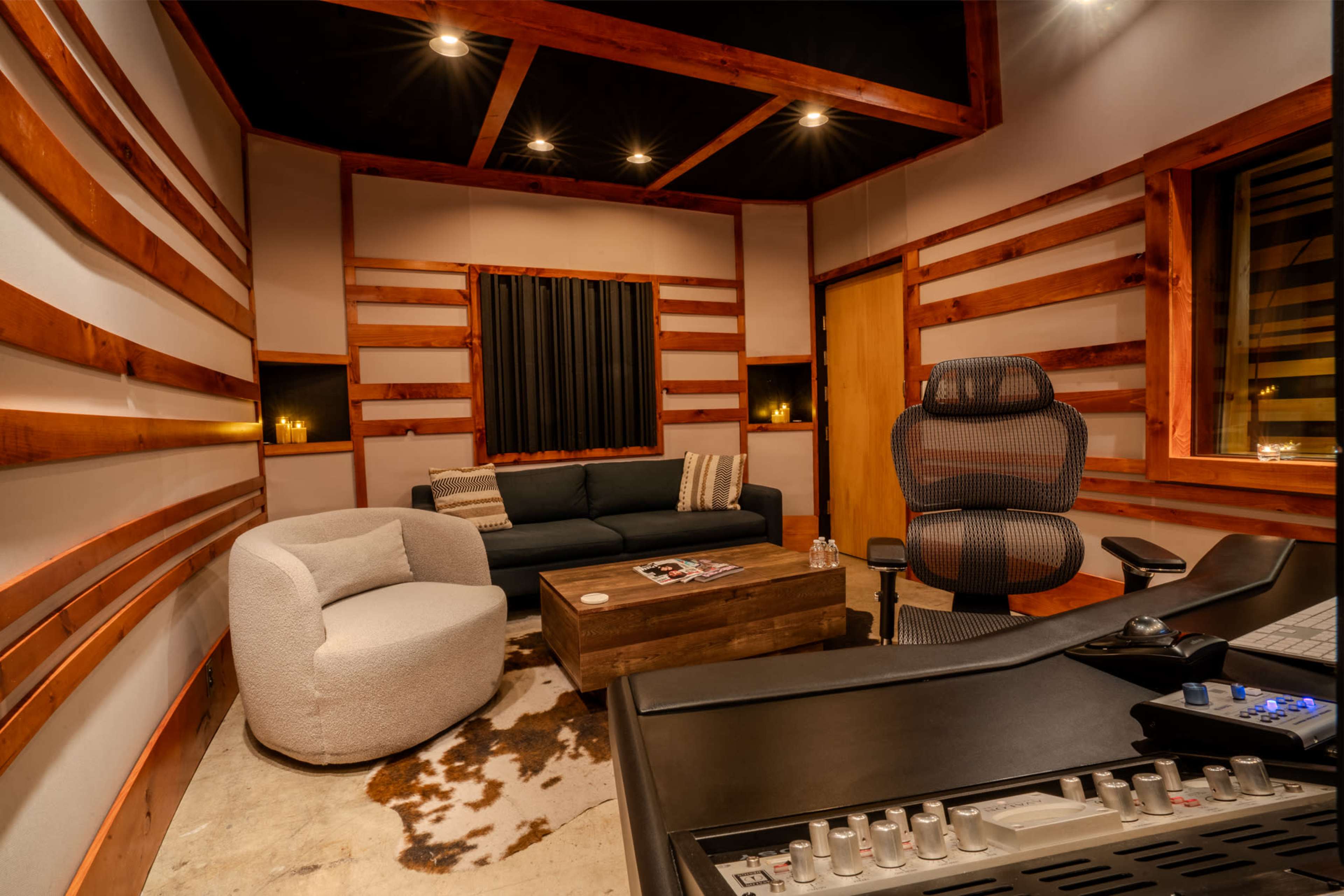 The image shows a modern recording studio featuring a dark wooden desk, a comfortable seating area with a sofa and an armchair, and acoustic panels on the walls.