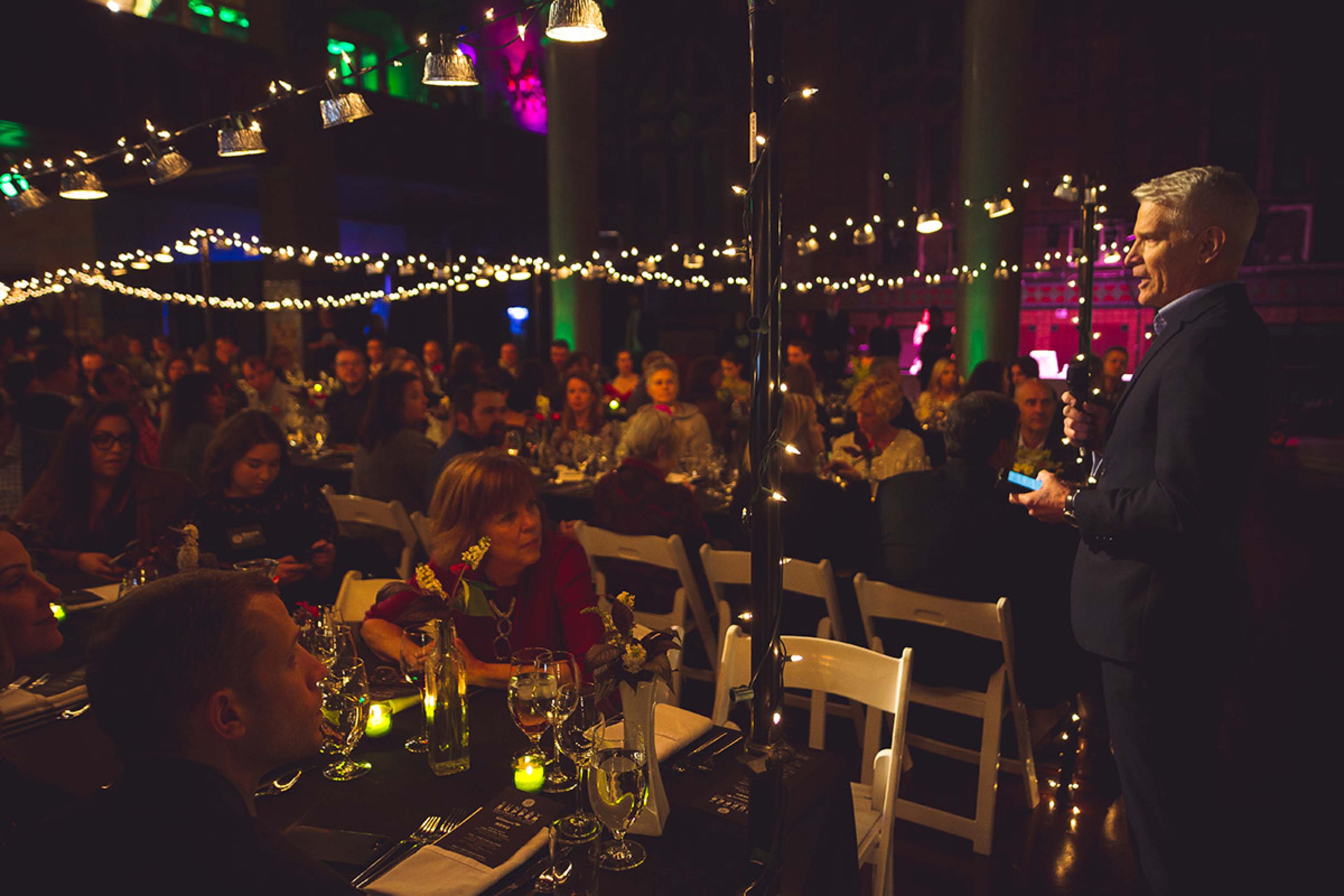 A speaker stands in front of a large audience seated at elegantly arranged tables illuminated by string lights and candles.