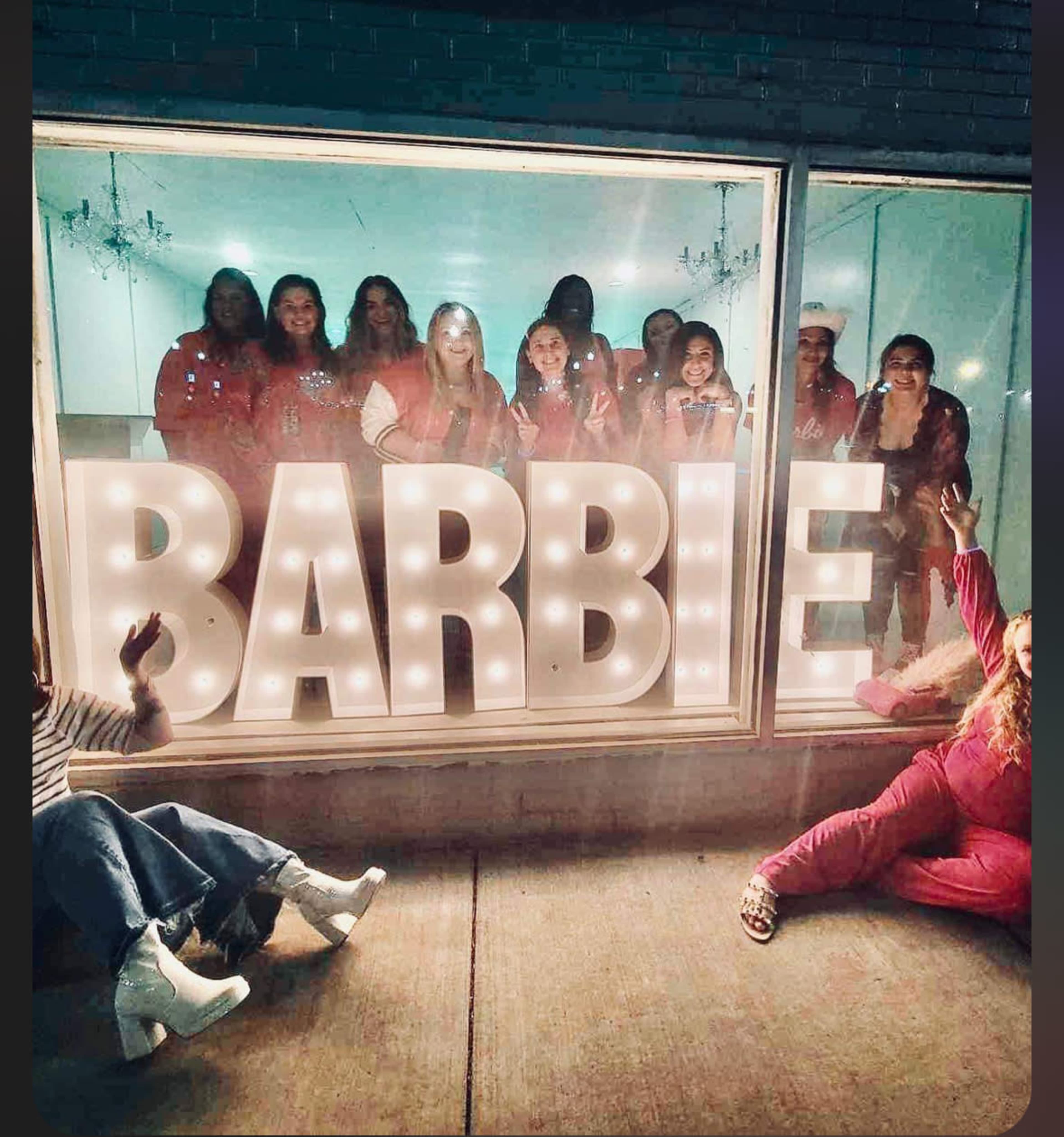 A group of women poses together in a storefront window beside large illuminated letters spelling "BARBIE."