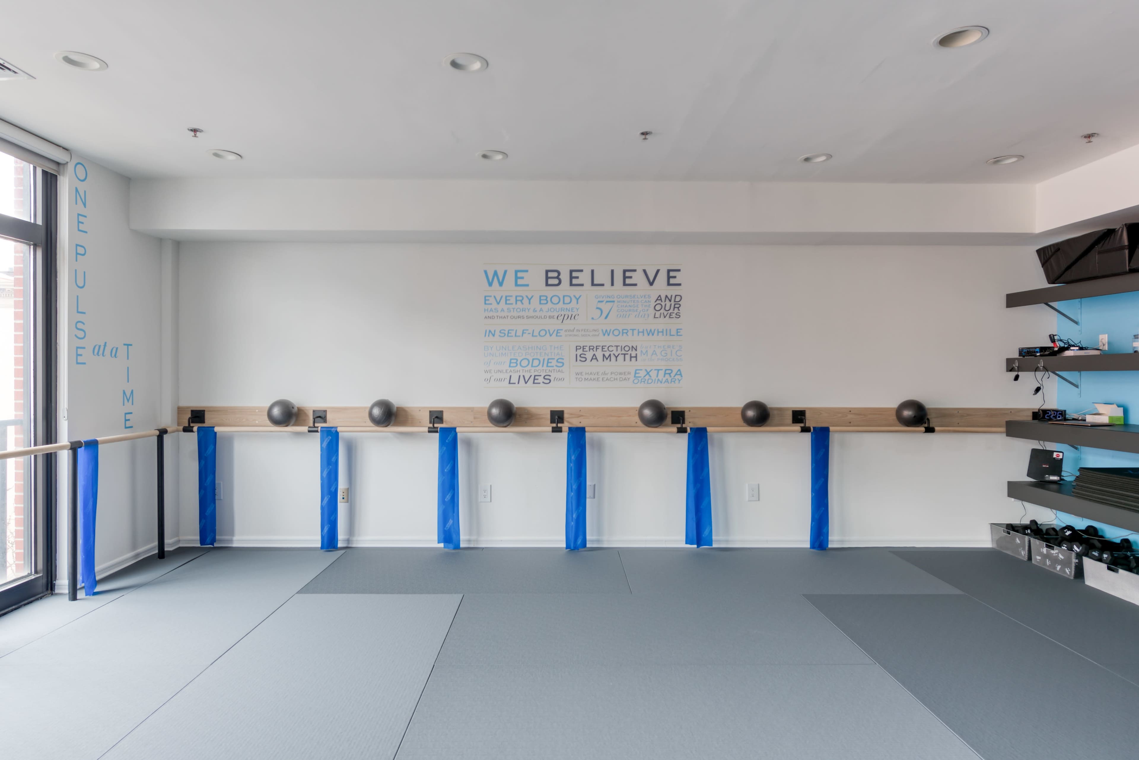 The image shows a fitness studio with blue exercise balls on wooden bars, gray mats on the floor, and motivational text on the wall.