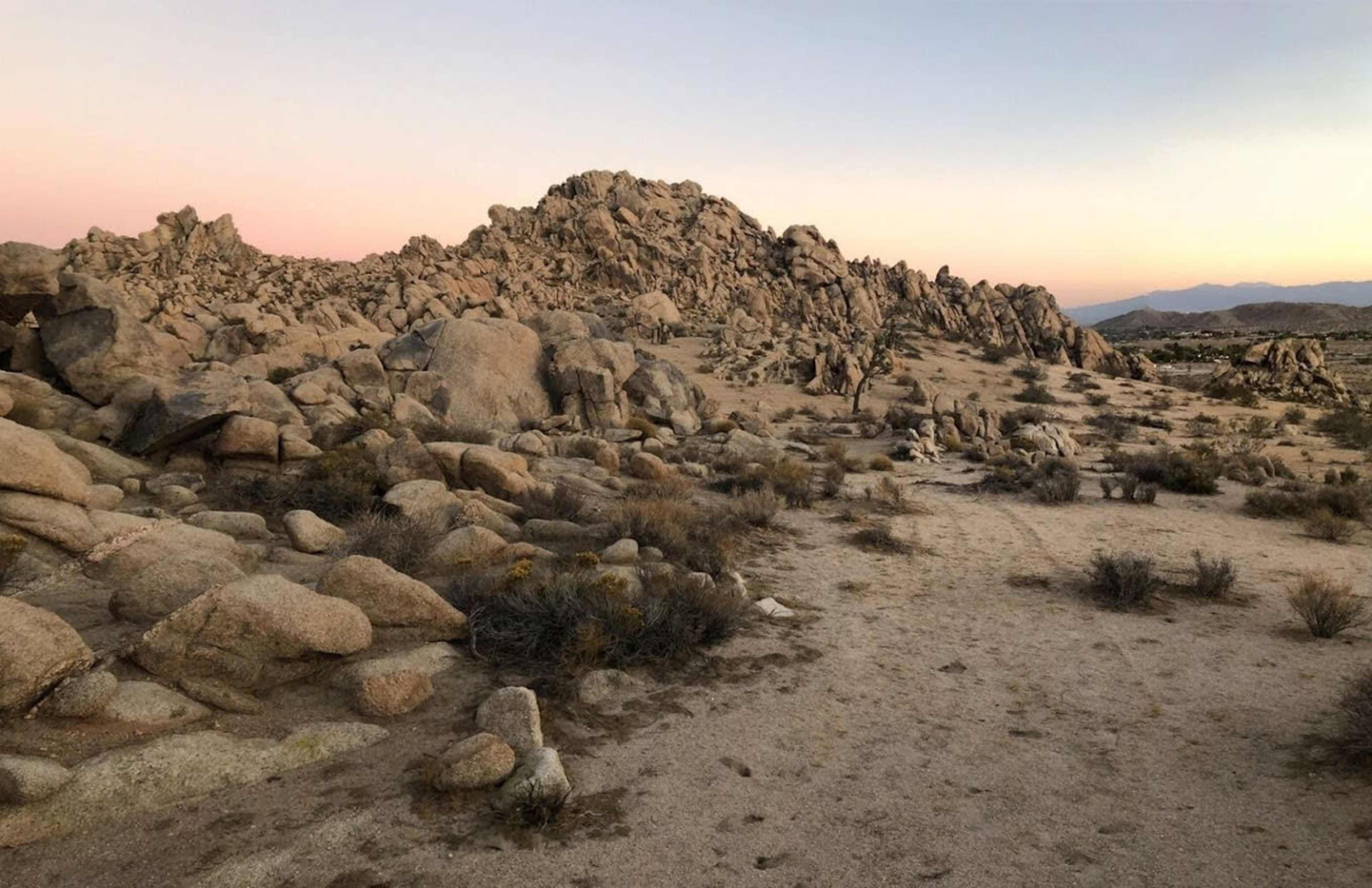 A rocky terrain is filled with boulders and sparse vegetation under a soft pastel sky at dusk.