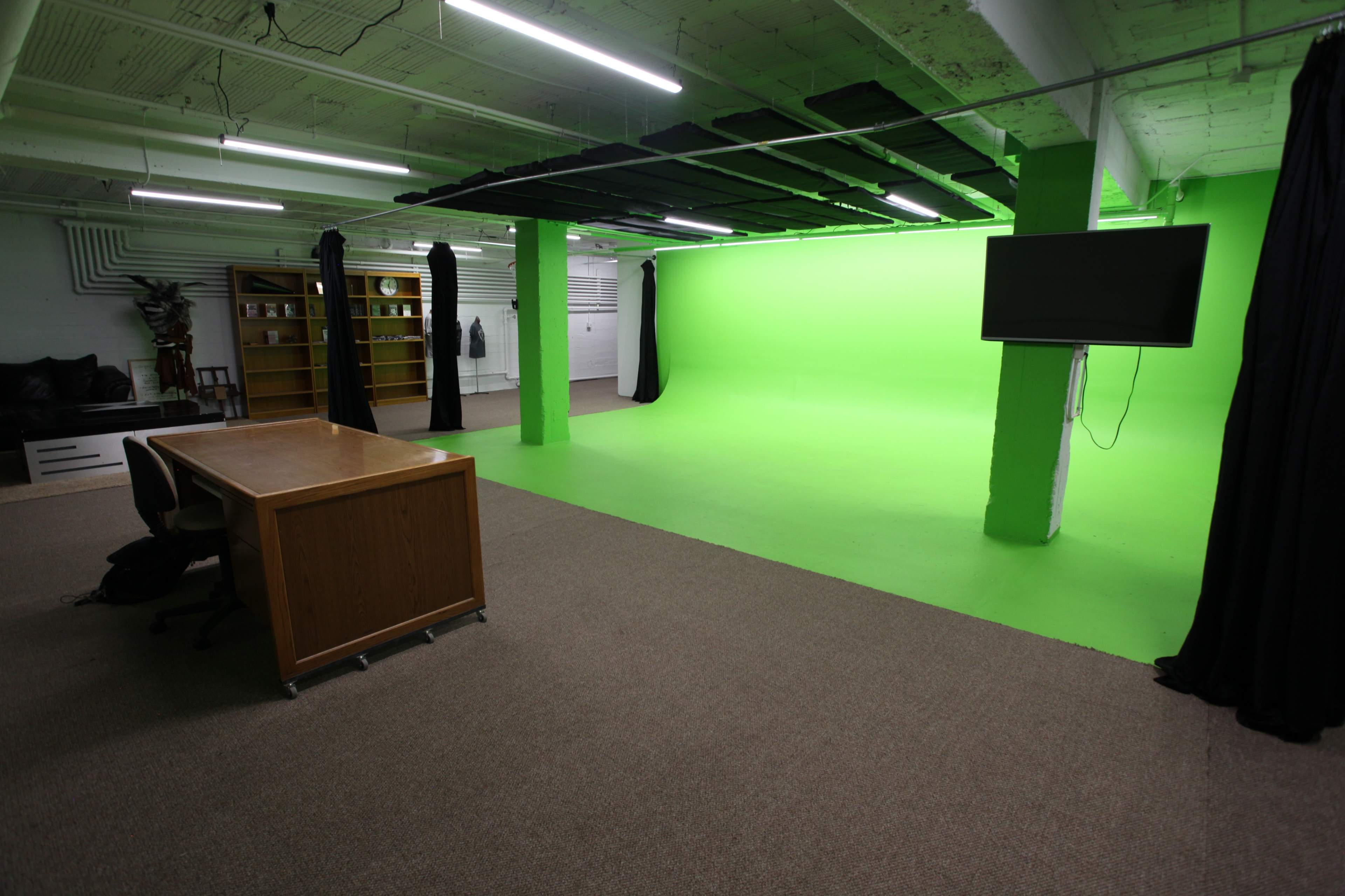 An indoor studio space featuring a green screen backdrop, a wooden desk with a chair, and shelves filled with various items.