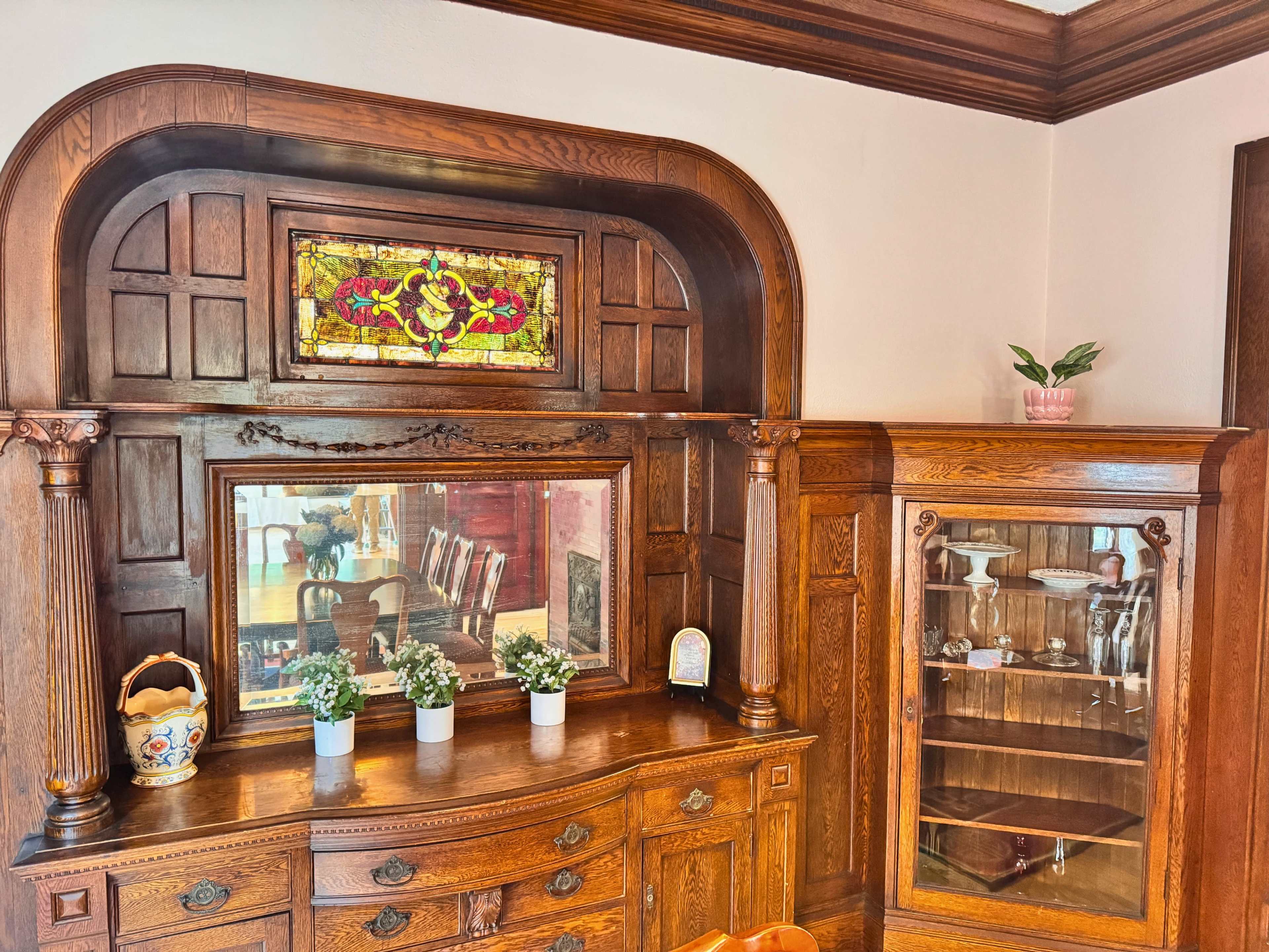 The image shows an ornate wooden sideboard with a stained glass window above it, flanked by a glass cabinet and decorative plants.