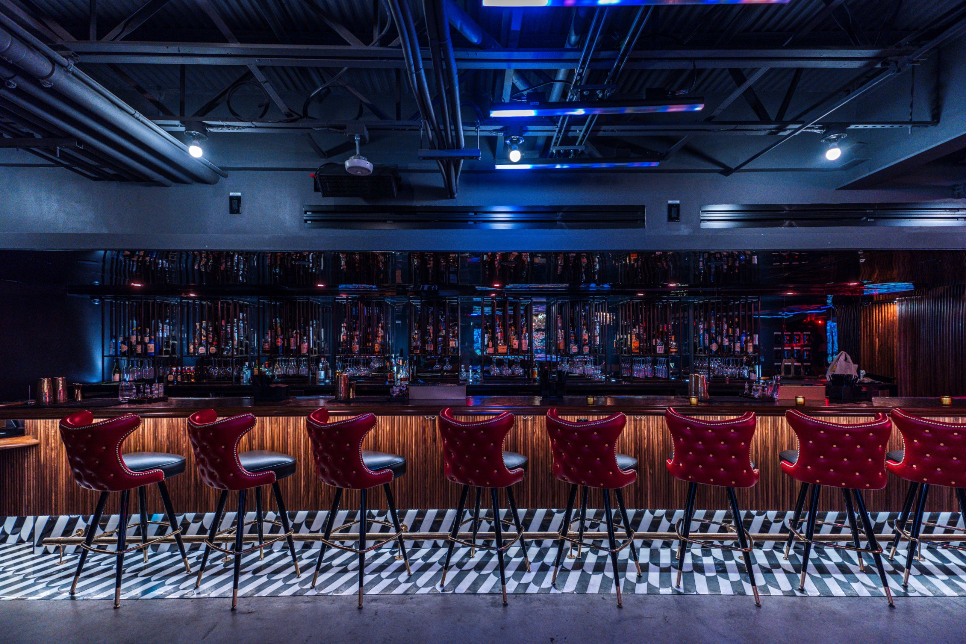 A modern bar area features a sleek design with red upholstered stools lined up against a wooden counter and illuminated by colorful lights.