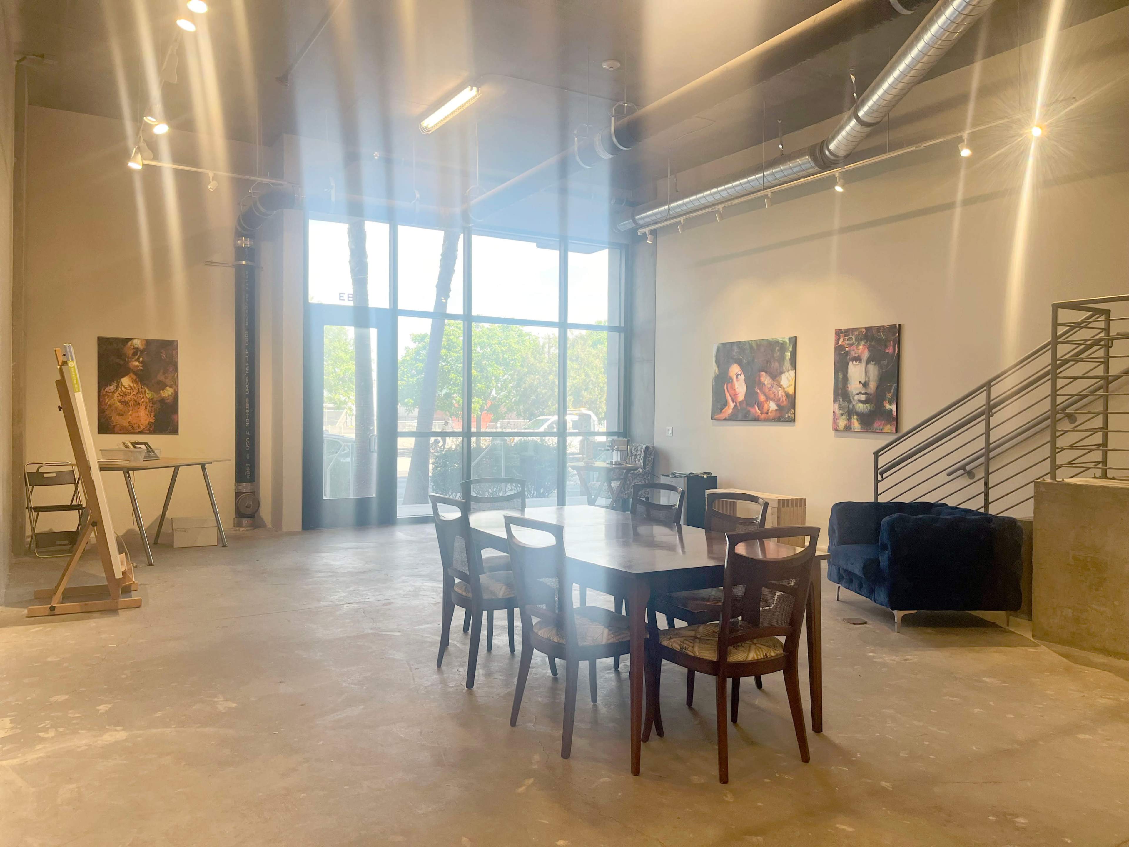 The image shows an art gallery with a wooden table surrounded by chairs, an easel, and large windows allowing natural light to enter, along with several framed artworks on the walls.