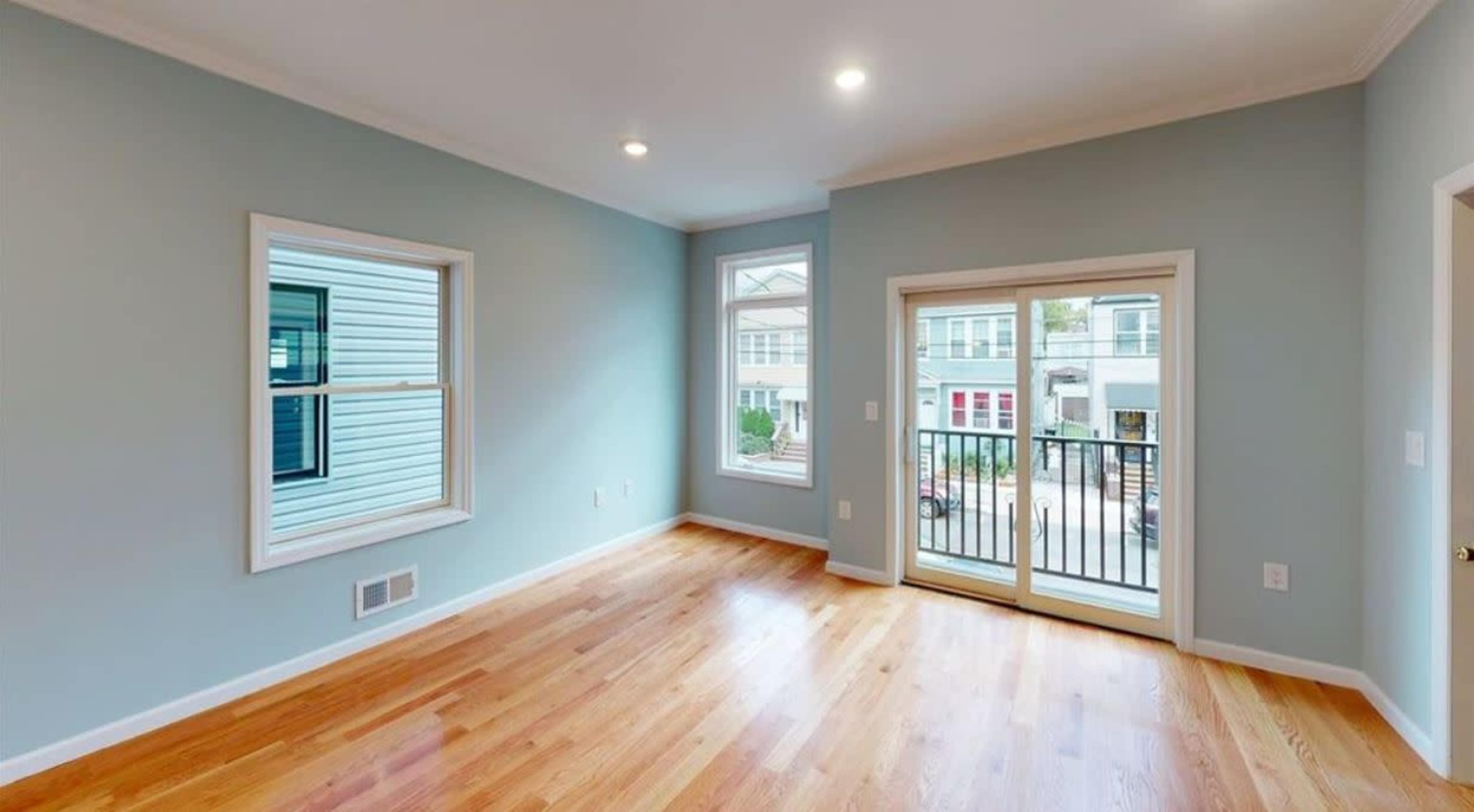 A spacious, light blue room with hardwood flooring and large windows, including a sliding door leading to a small balcony.