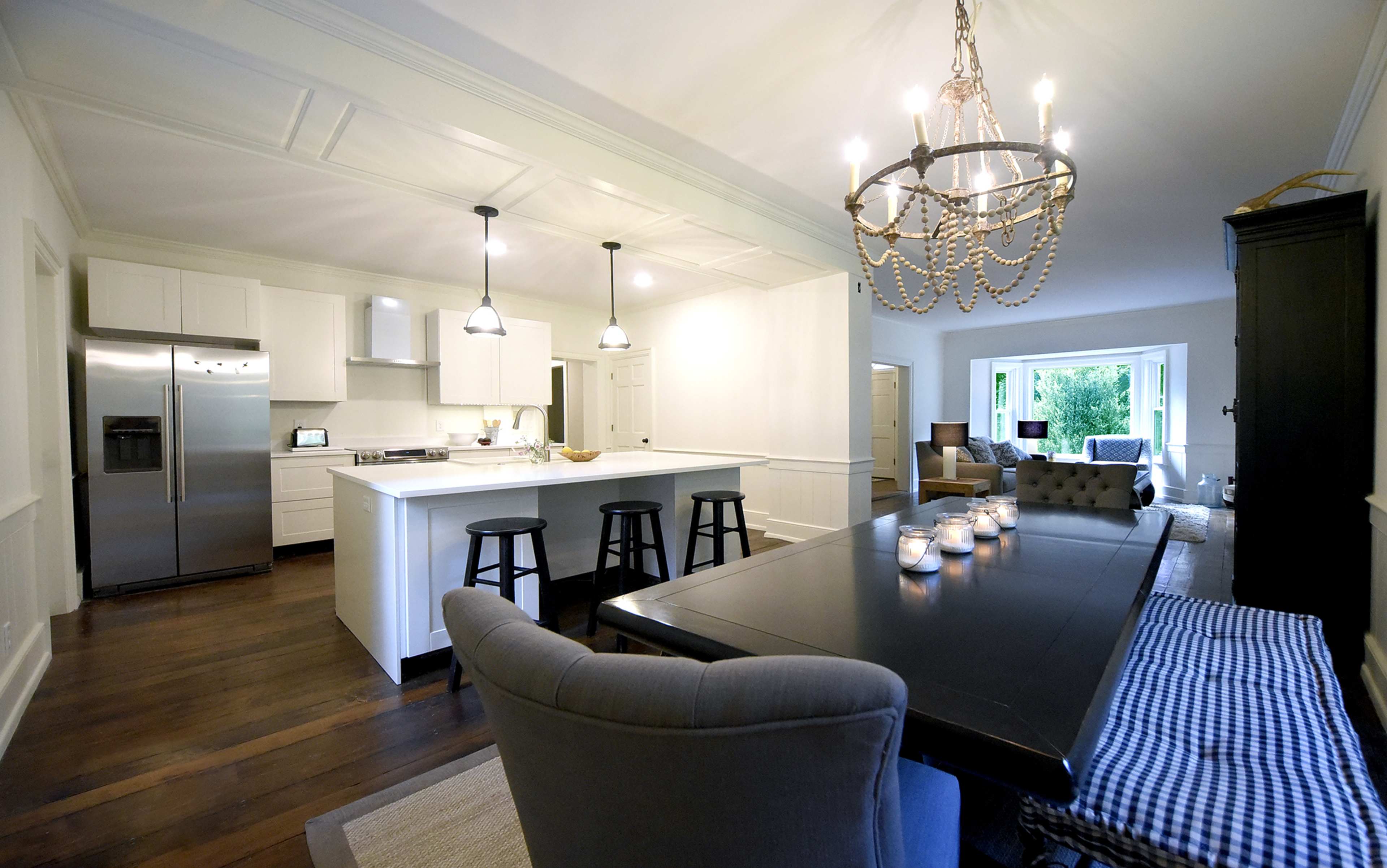 A modern kitchen with a large dining table, black stools at the island, and a chandelier overhead, set in a bright room with wood flooring.