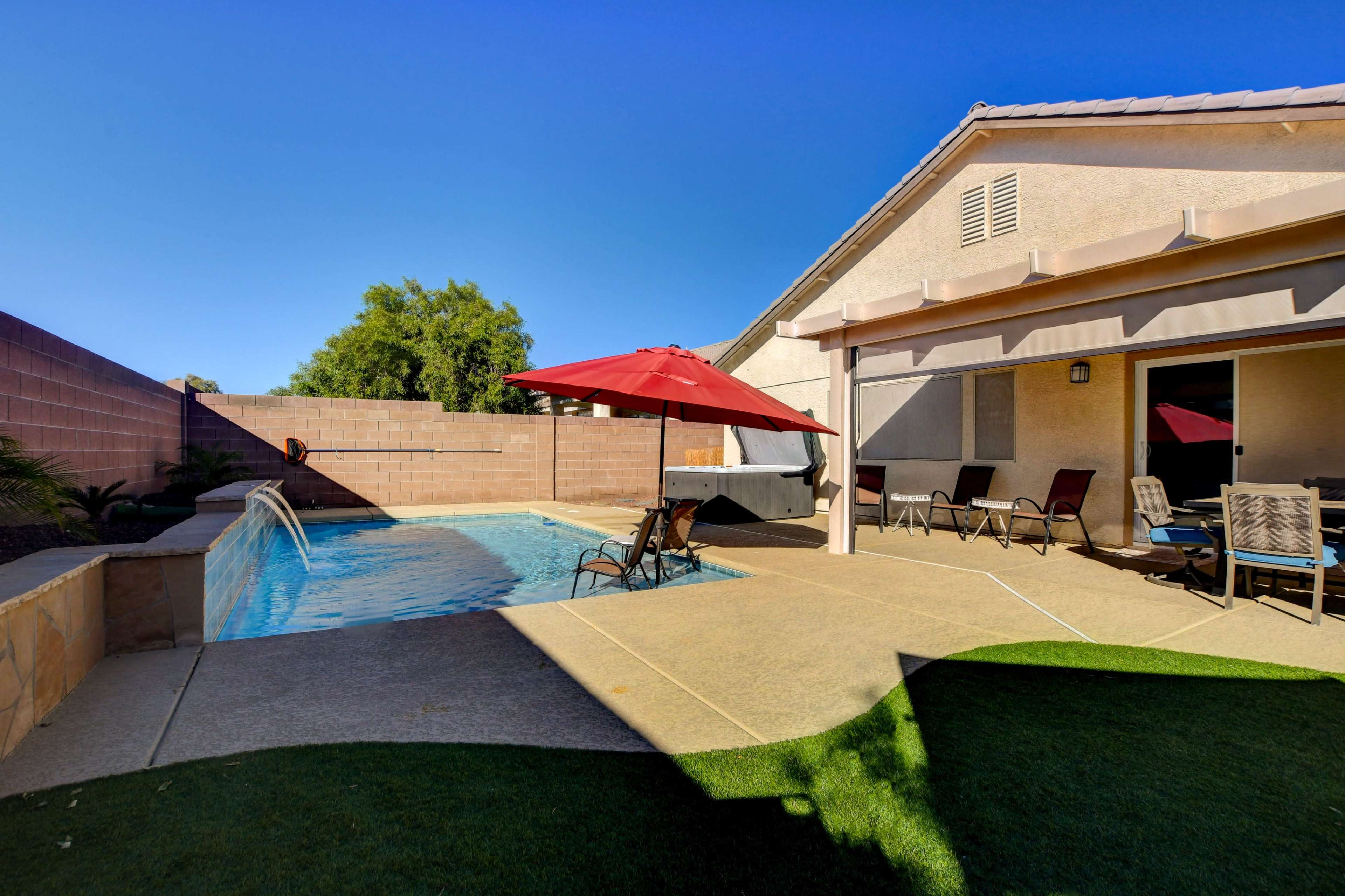 A backyard with a swimming pool, a red umbrella, lounge chairs, and a beige house under a clear blue sky.