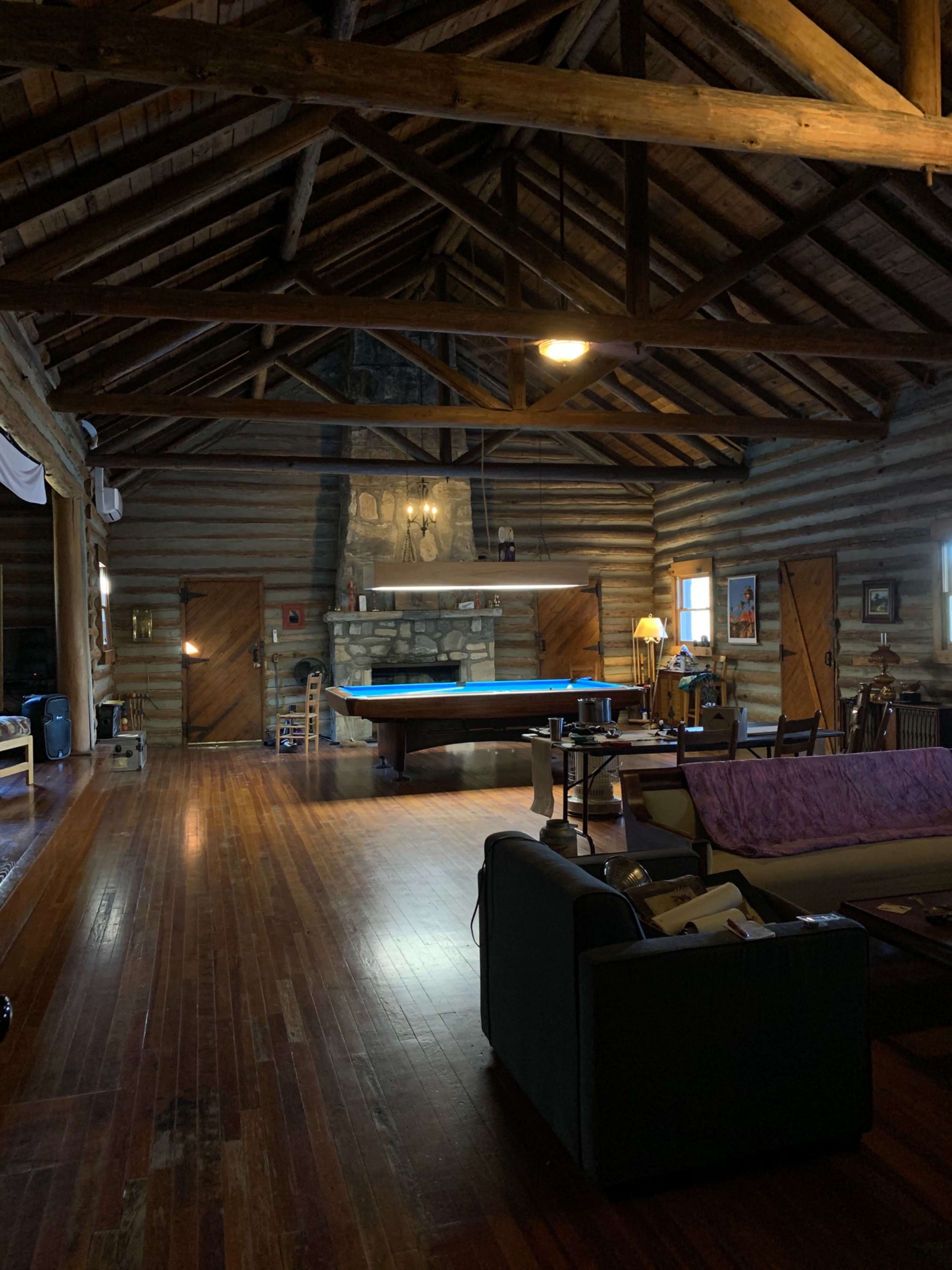 The image shows a spacious cabin interior with wooden beams and a pool table in the center, surrounded by various furniture and a stone fireplace.