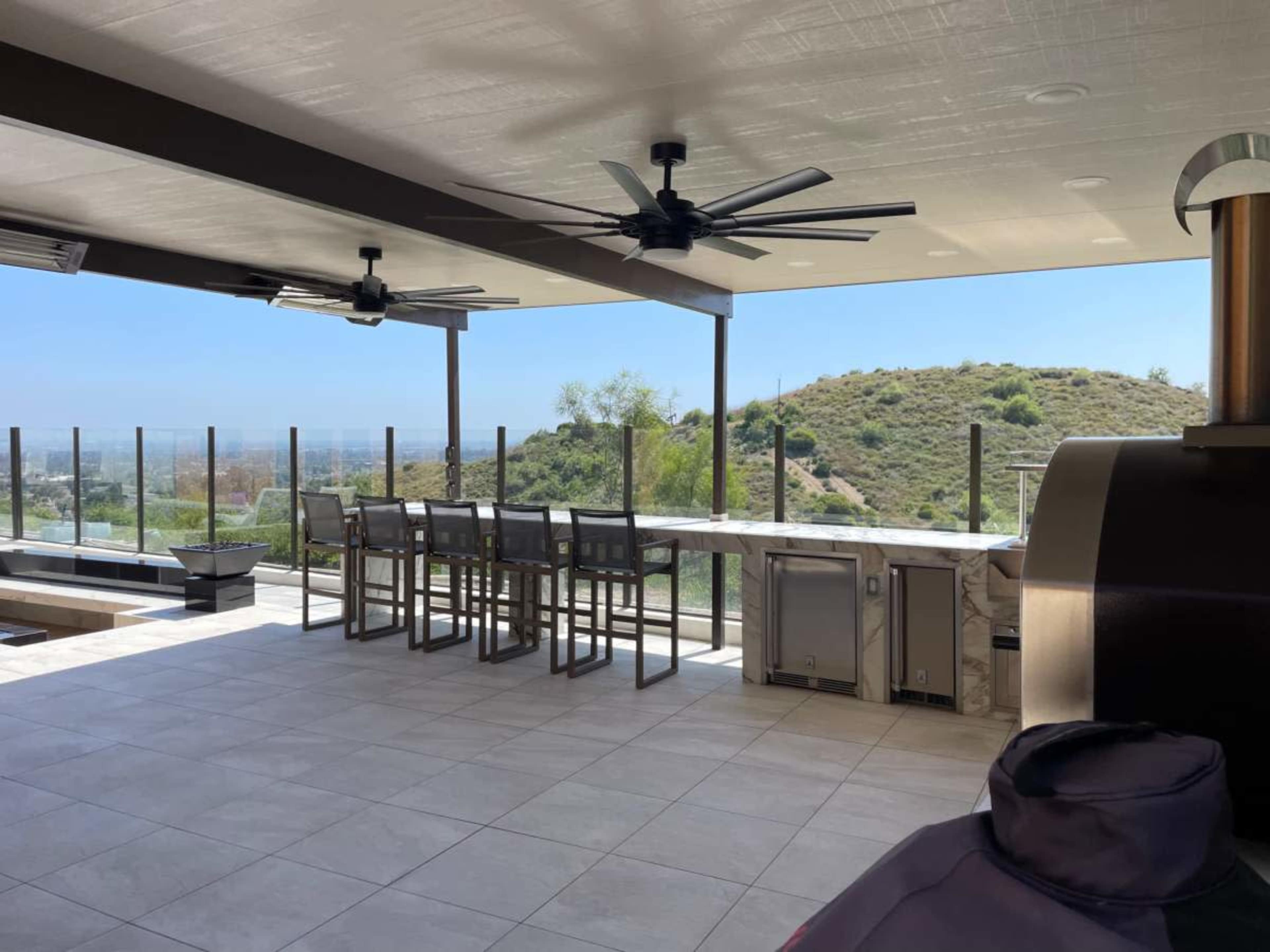 An open outdoor kitchen area with several bar stools, a countertop, and a view of a hillside.