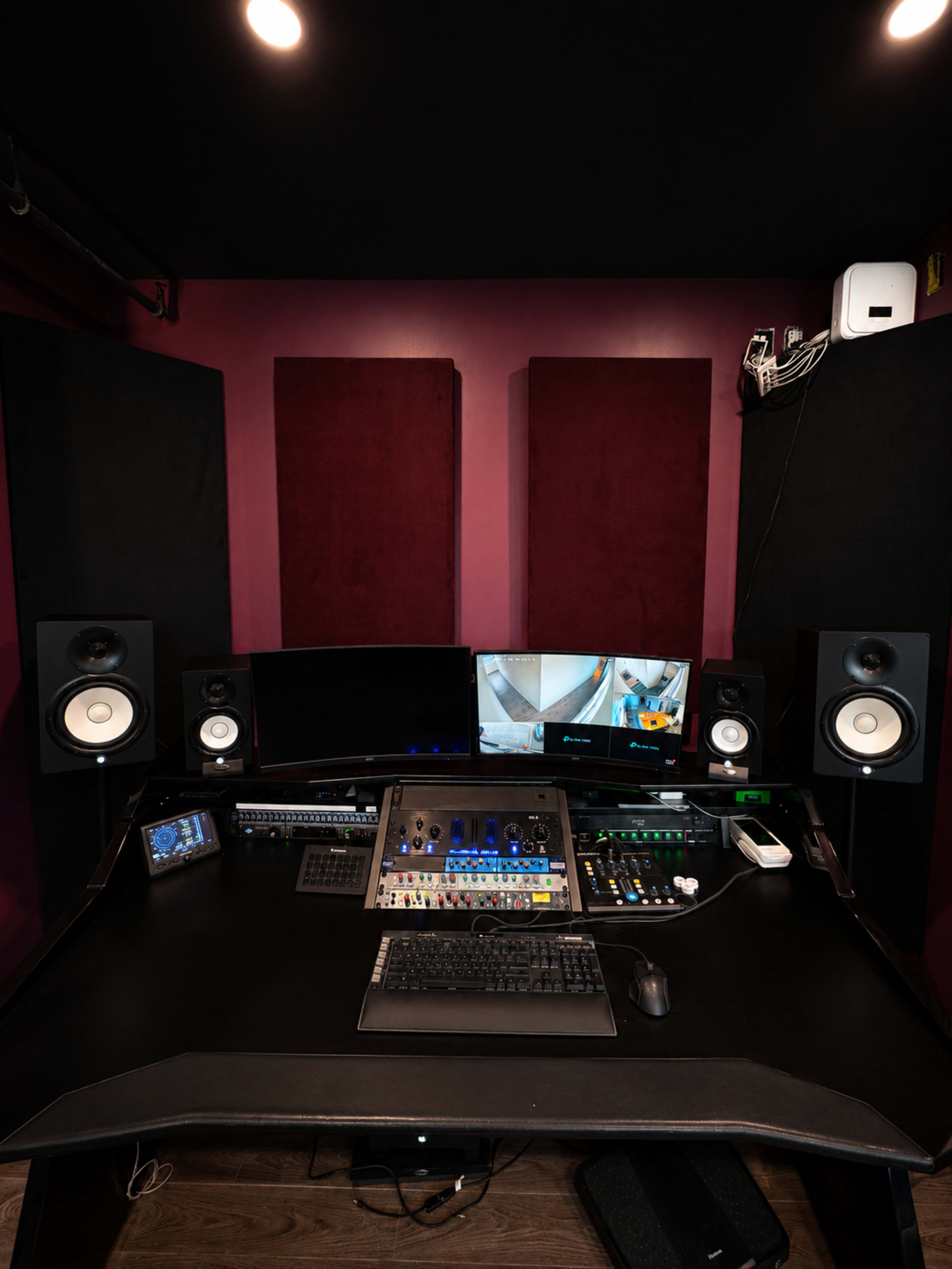 The image shows a music production studio featuring a black desk with audio equipment, two monitors, speakers, and soundproofing panels on the walls.