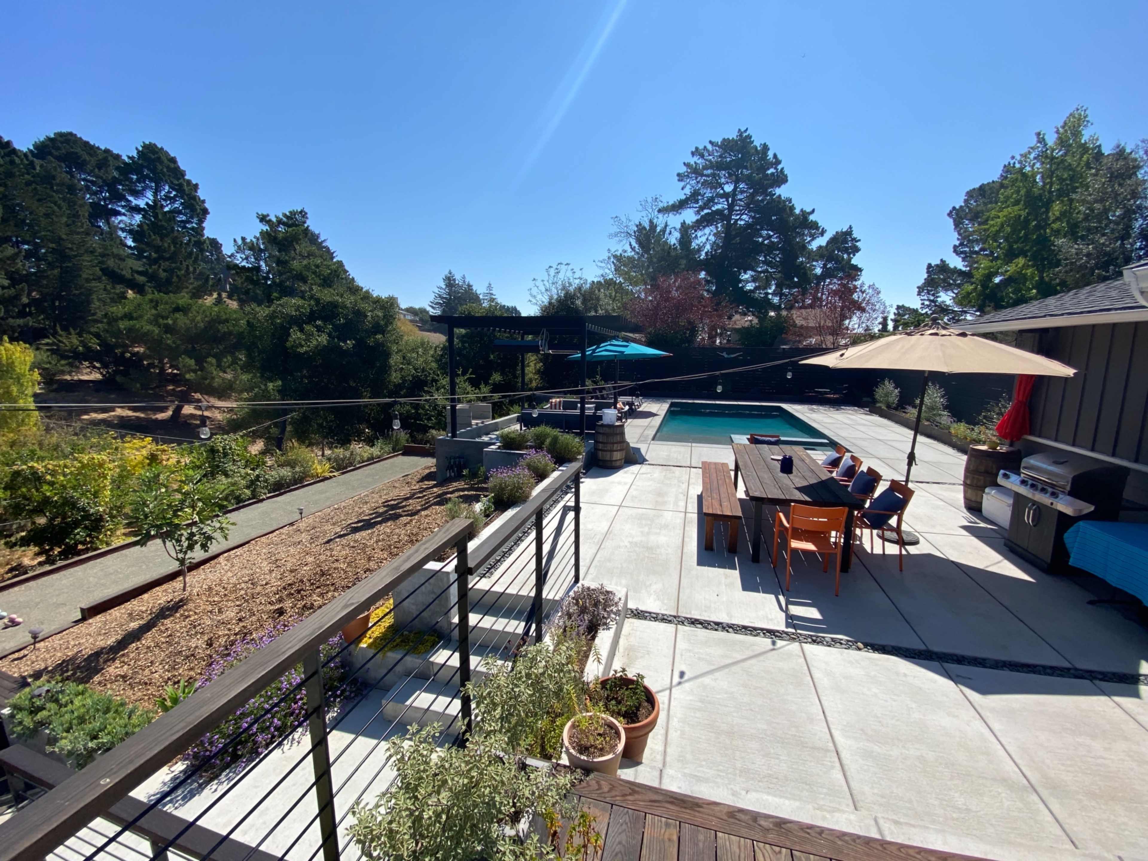 A backyard with a swimming pool, a dining table, and a barbeque grill, surrounded by landscaping and trees under a clear blue sky.