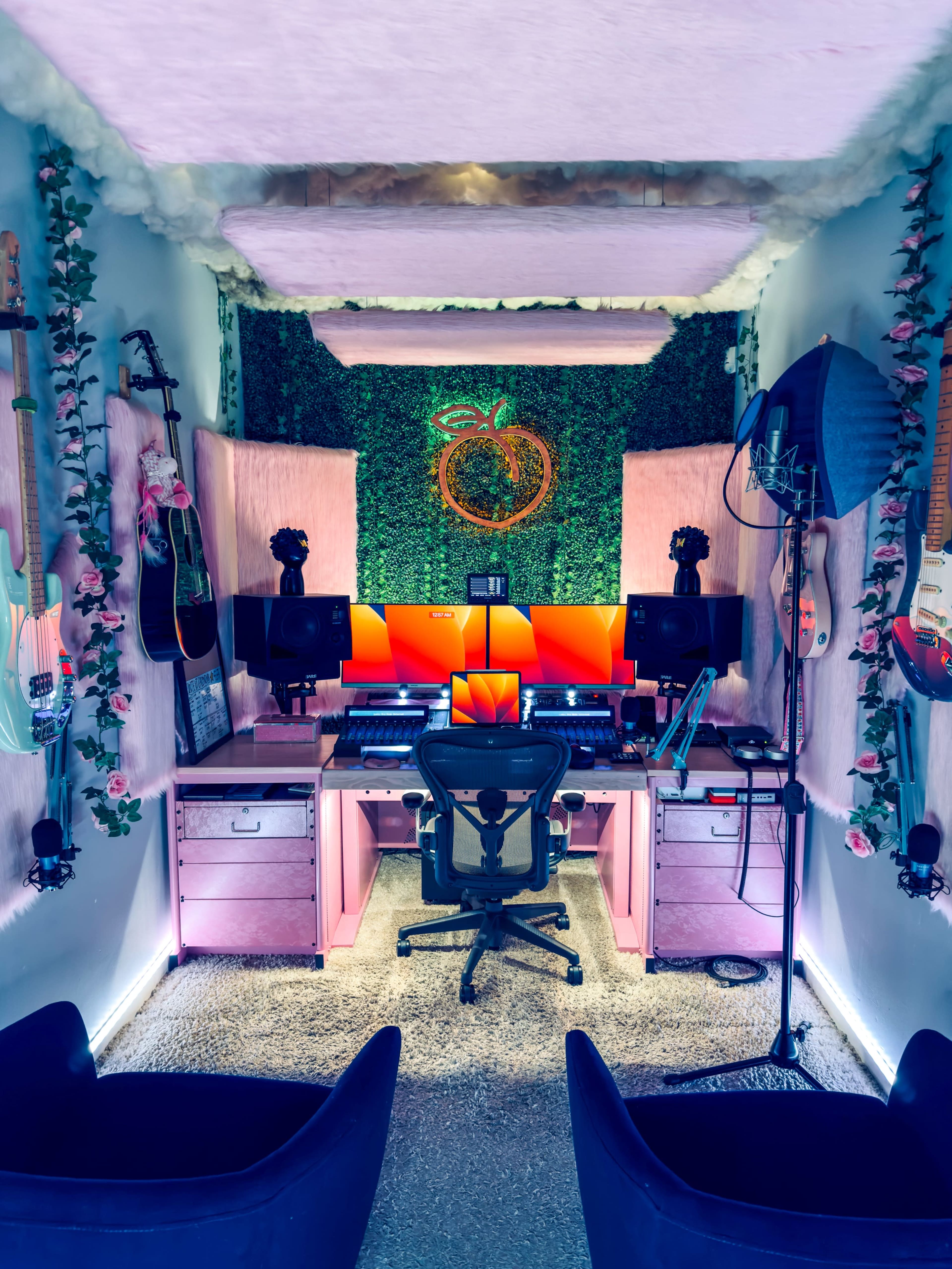A music studio featuring two computer workstations, surrounded by plants and musical instruments on the walls.