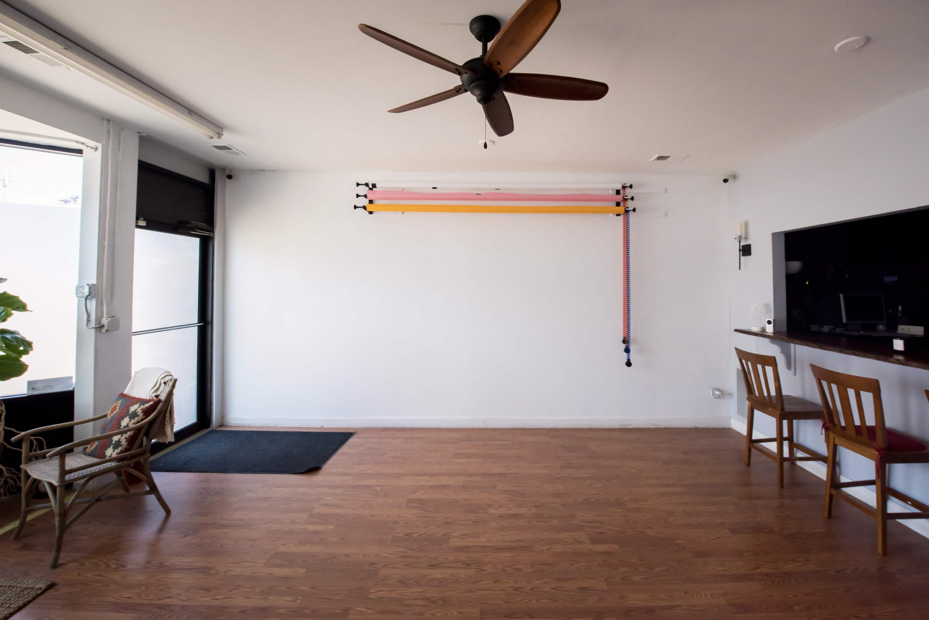 A spacious, sparsely furnished room features a ceiling fan, a bar with two chairs, and exercise bands mounted on the wall.