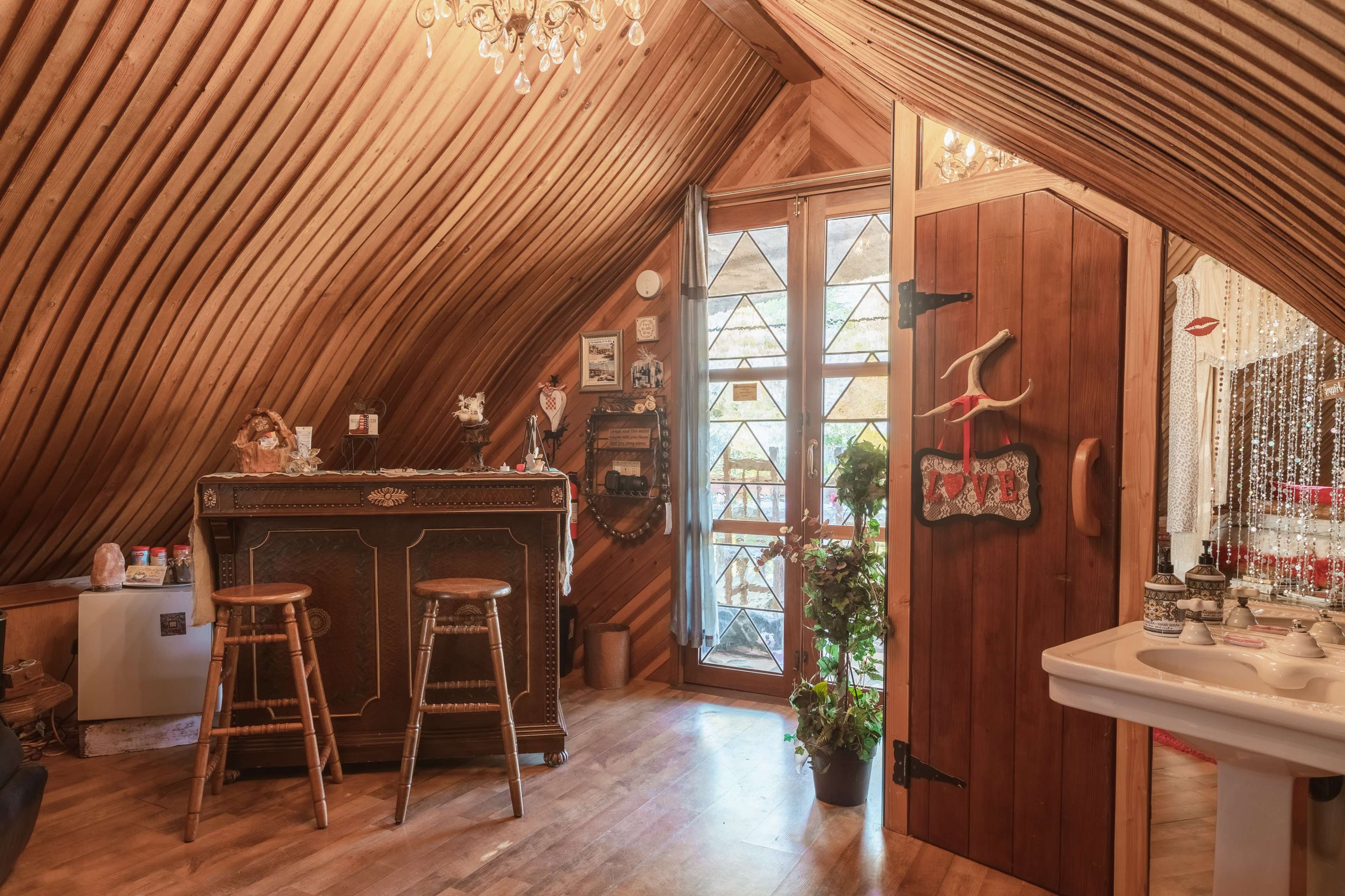 A cozy attic room with a wooden ceiling, a bar-style counter with two stools, and a glass door leading to an outdoor space.