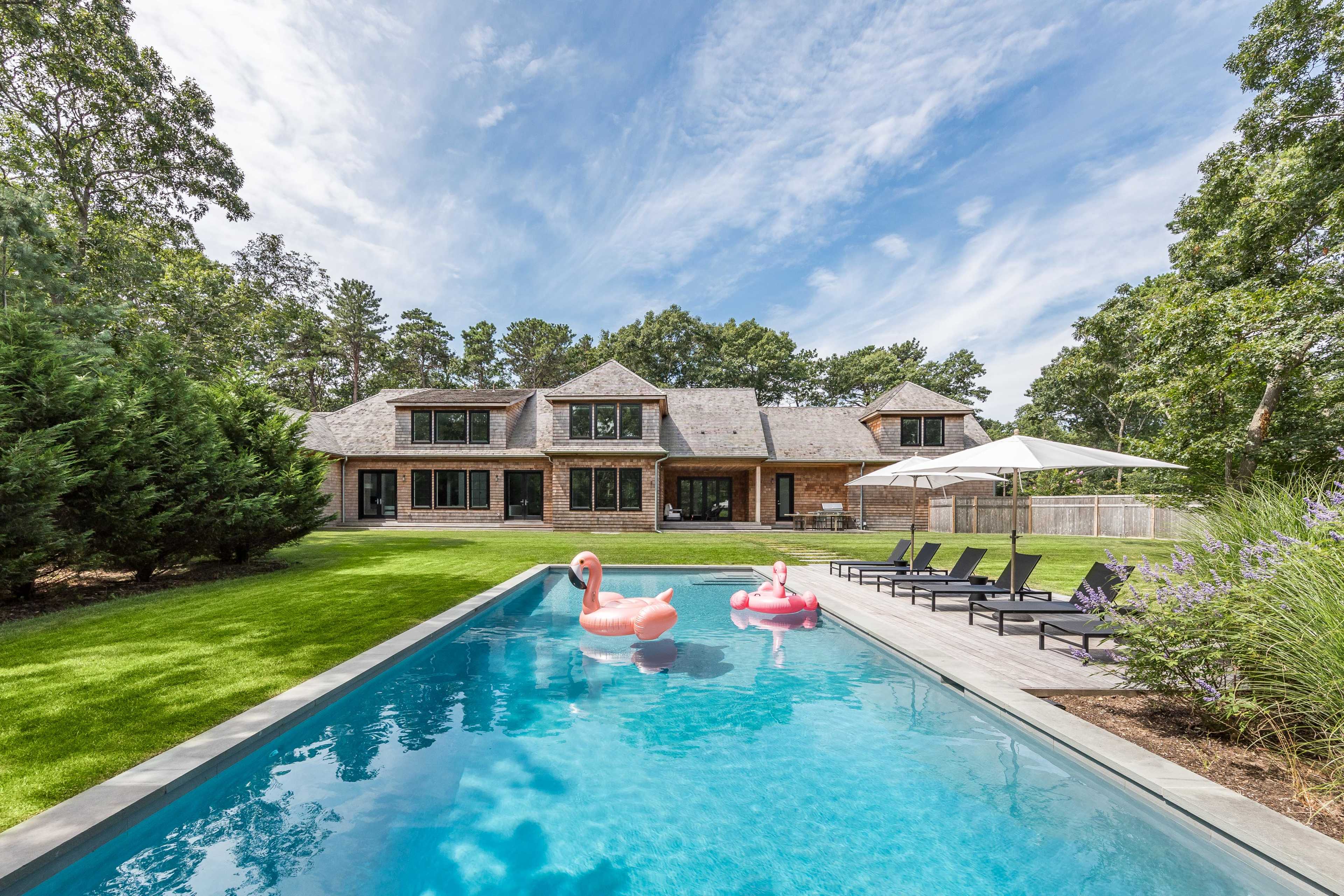 A modern house with a shingle roof is situated next to a swimming pool featuring pink flamingo floats, surrounded by a well-maintained lawn and lounging chairs.