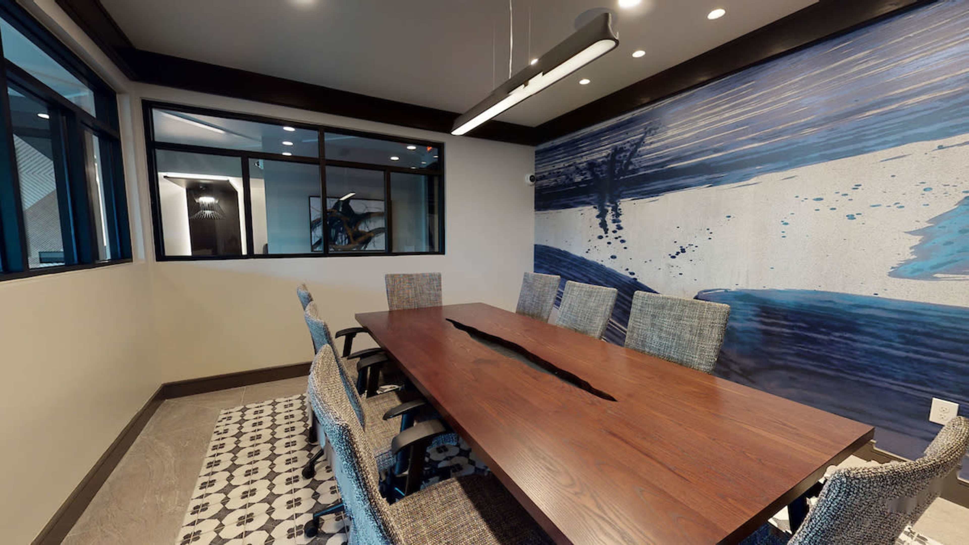 A conference room features a long wooden table surrounded by upholstered chairs, with a large blue mural on one wall and floor-to-ceiling windows providing natural light.