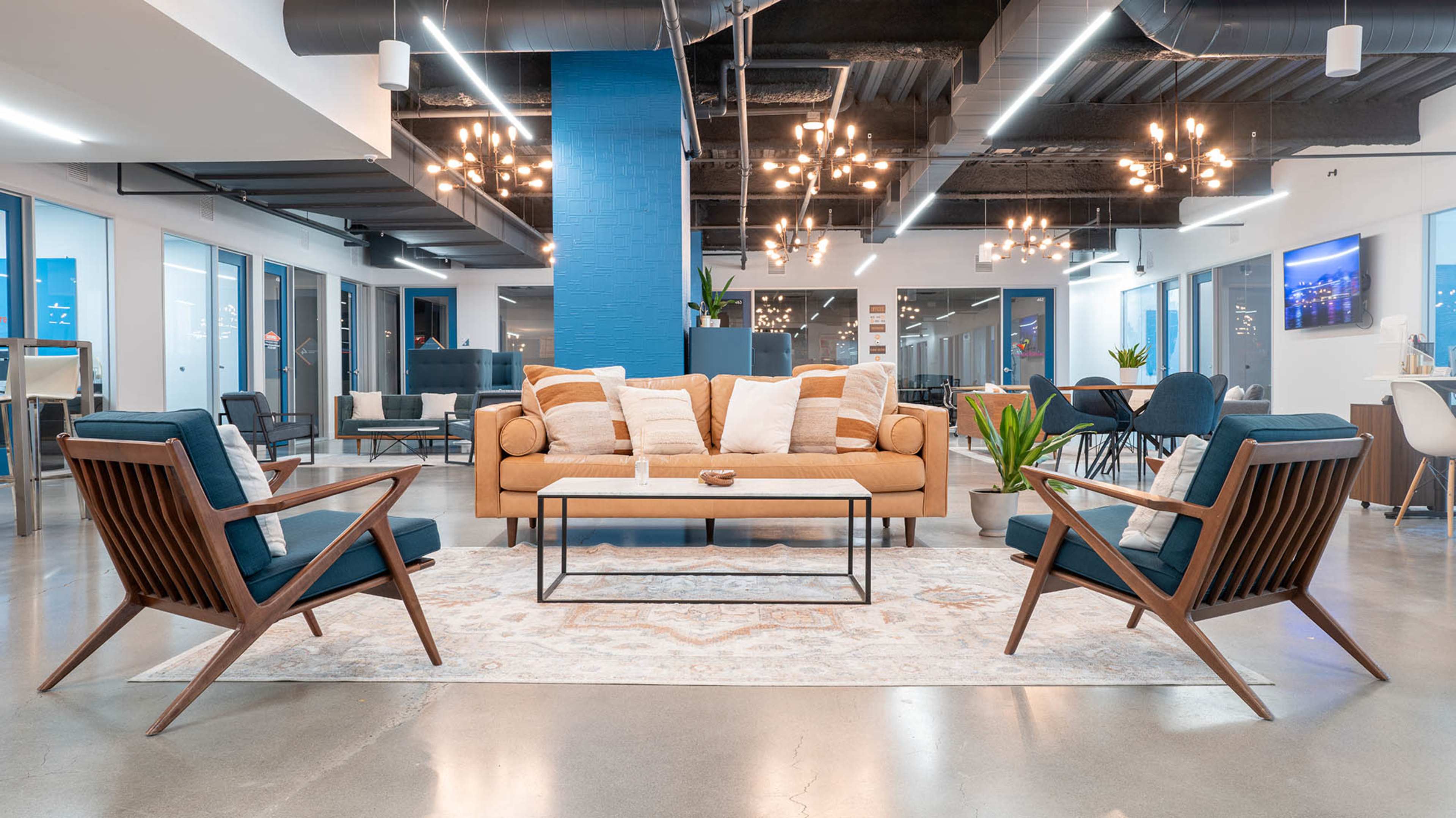 The image shows a modern office lounge area featuring a tan sofa, two dark blue accent chairs, a coffee table, and various plants under decorative ceiling lights.