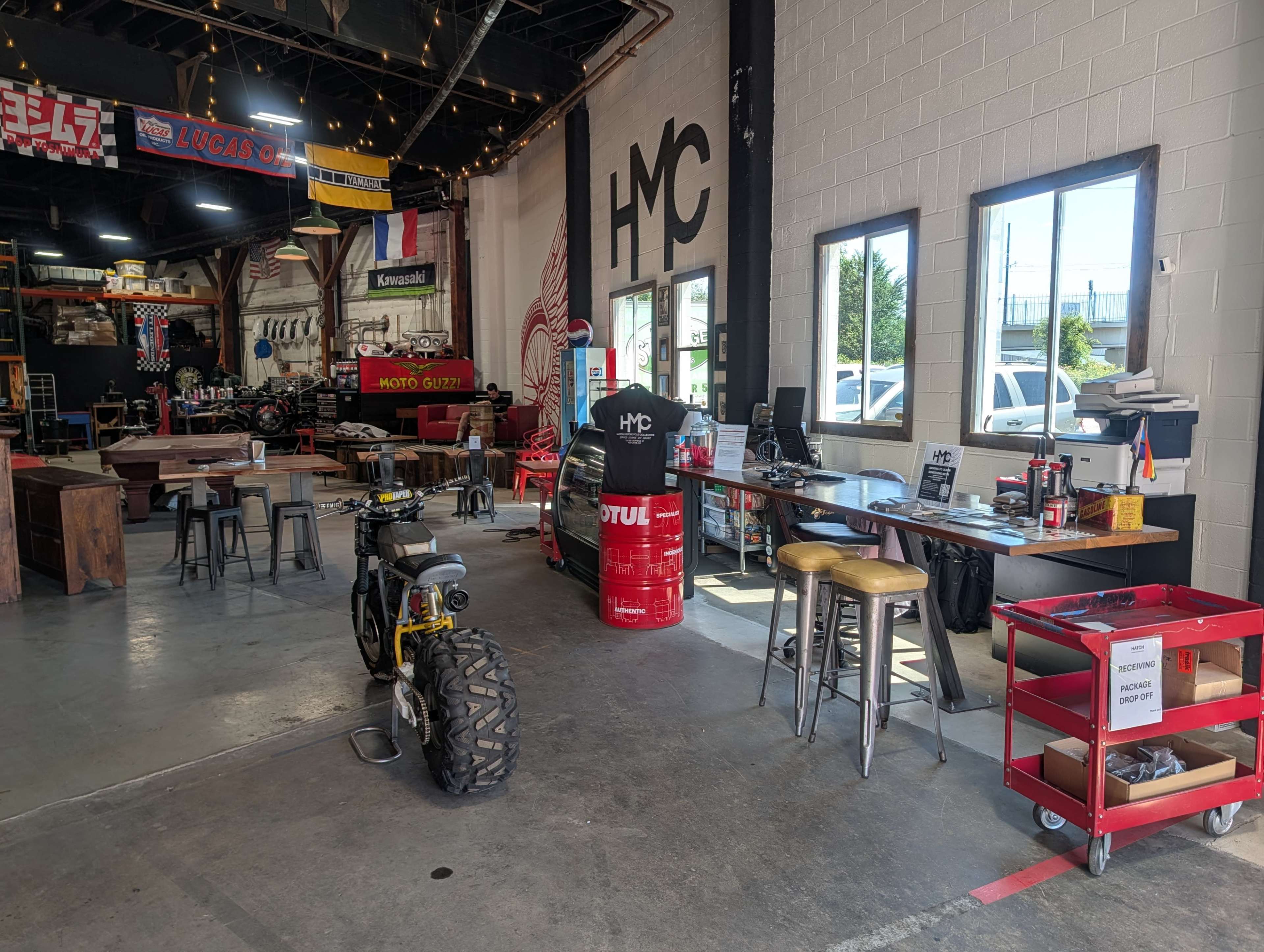 The interior of a motorcycle workshop features workstations, a vintage bike, and various tools, with flags and banners adorning the walls.