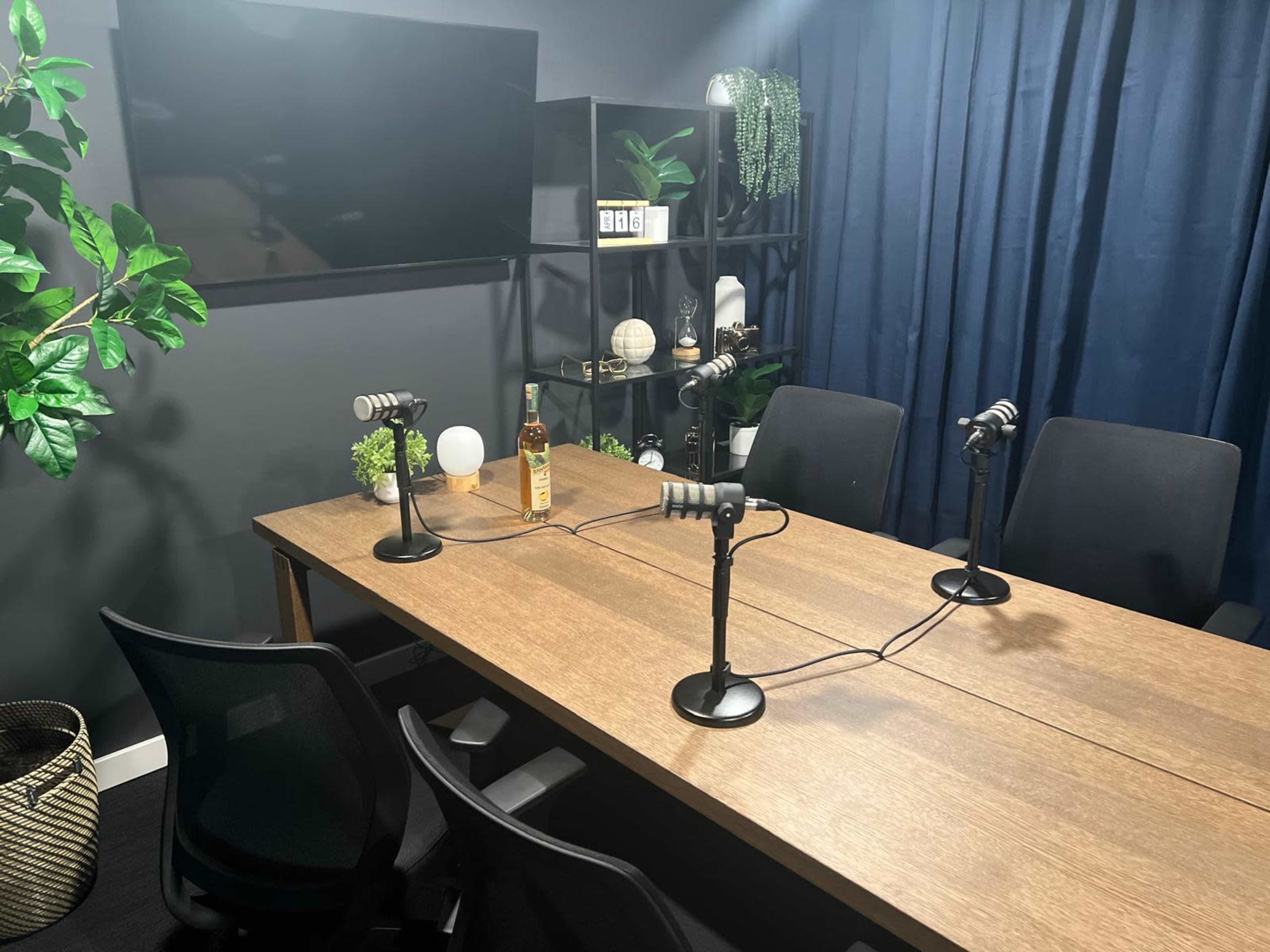 A conference room table with four microphones is set up for a podcast recording, surrounded by black chairs and decorated with plants and shelves.