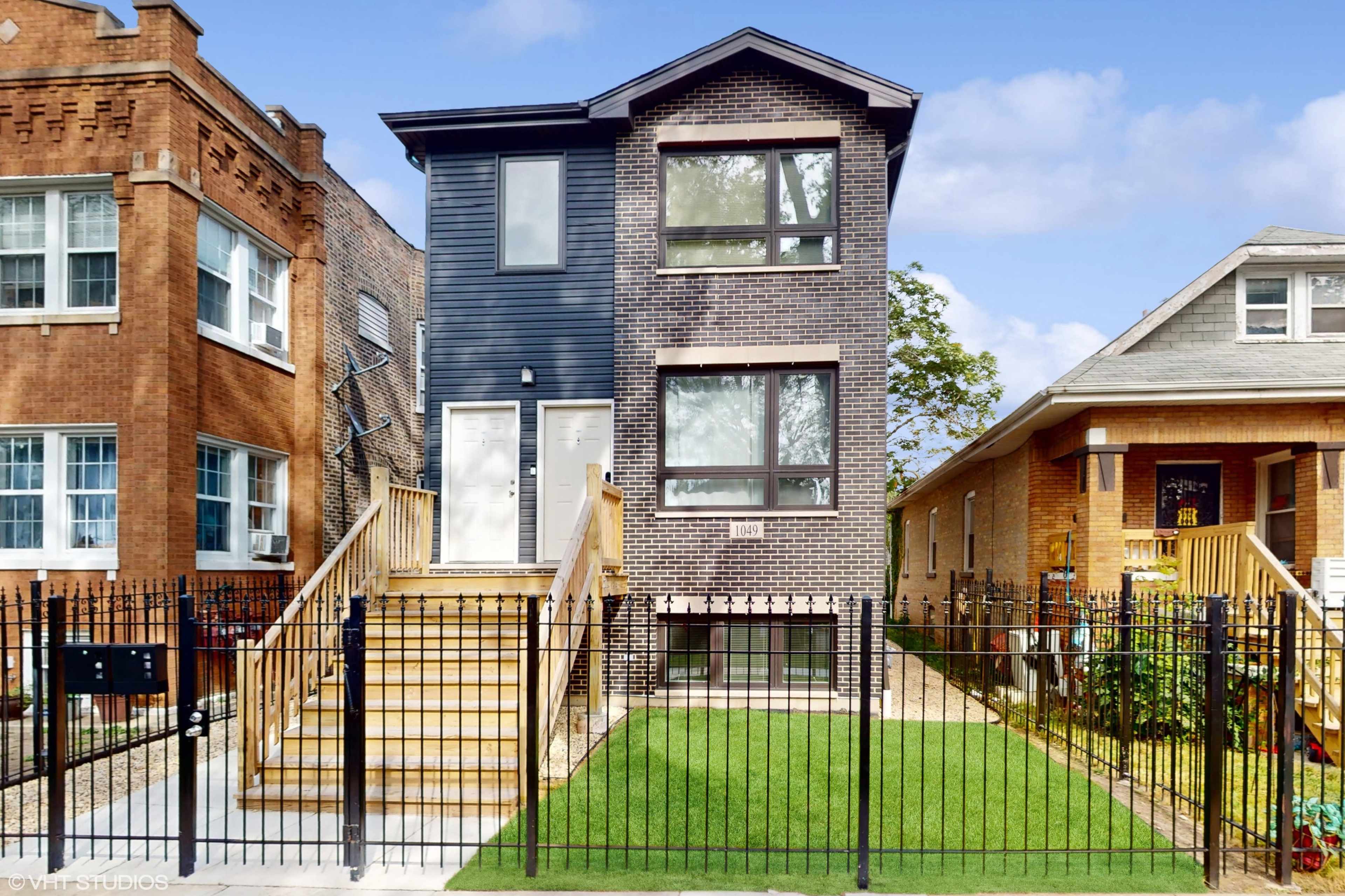 A modern two-story residential building with a black and gray brick facade, flanked by two neighboring homes and featuring a fenced yard with green grass.
