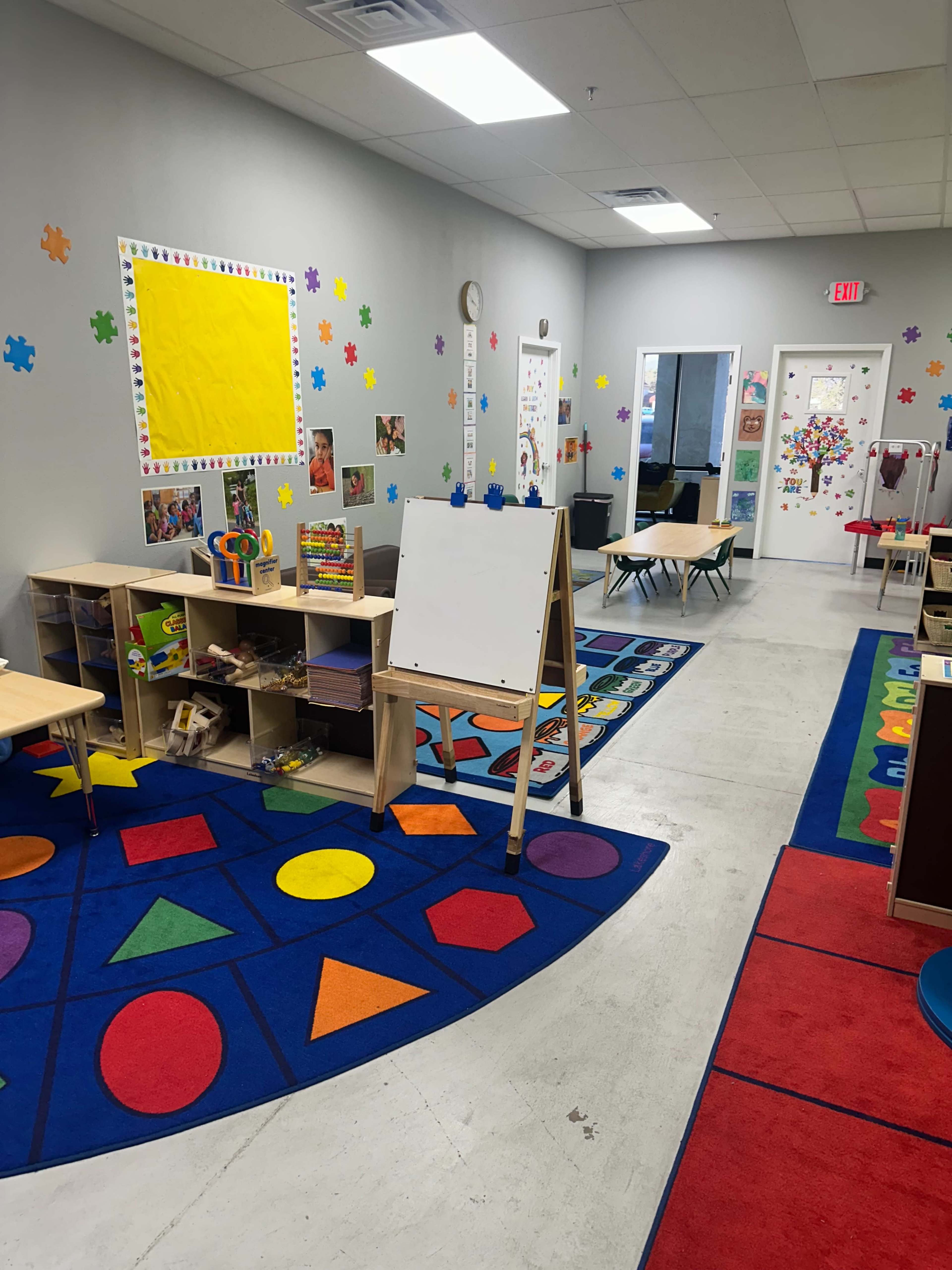 The image shows a colorful preschool classroom with a variety of educational materials, rugs featuring geometric shapes, and decorated walls.