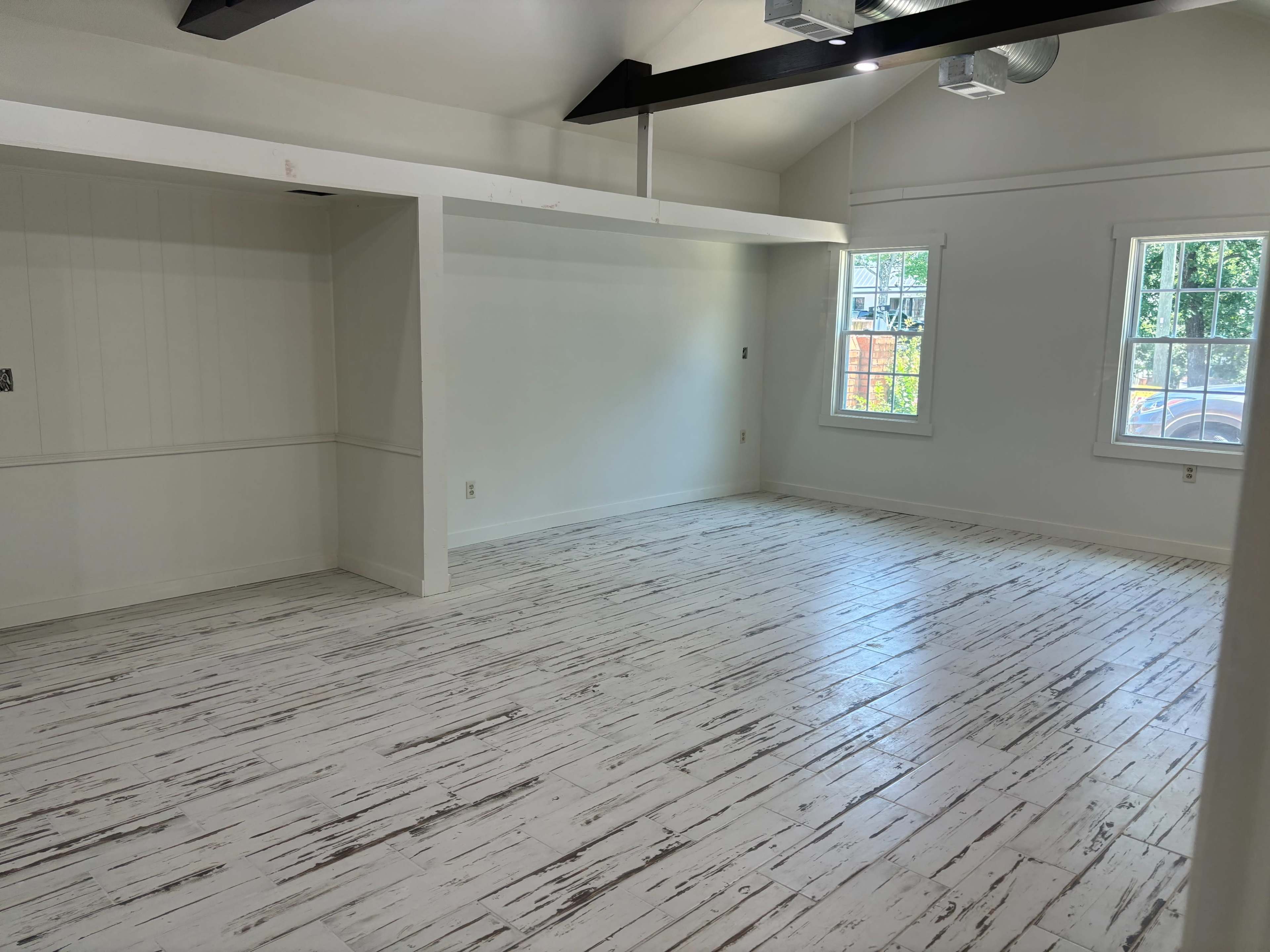 The image shows a spacious, empty interior room with white walls, a wood ceiling with black beams, and a weathered white floor.