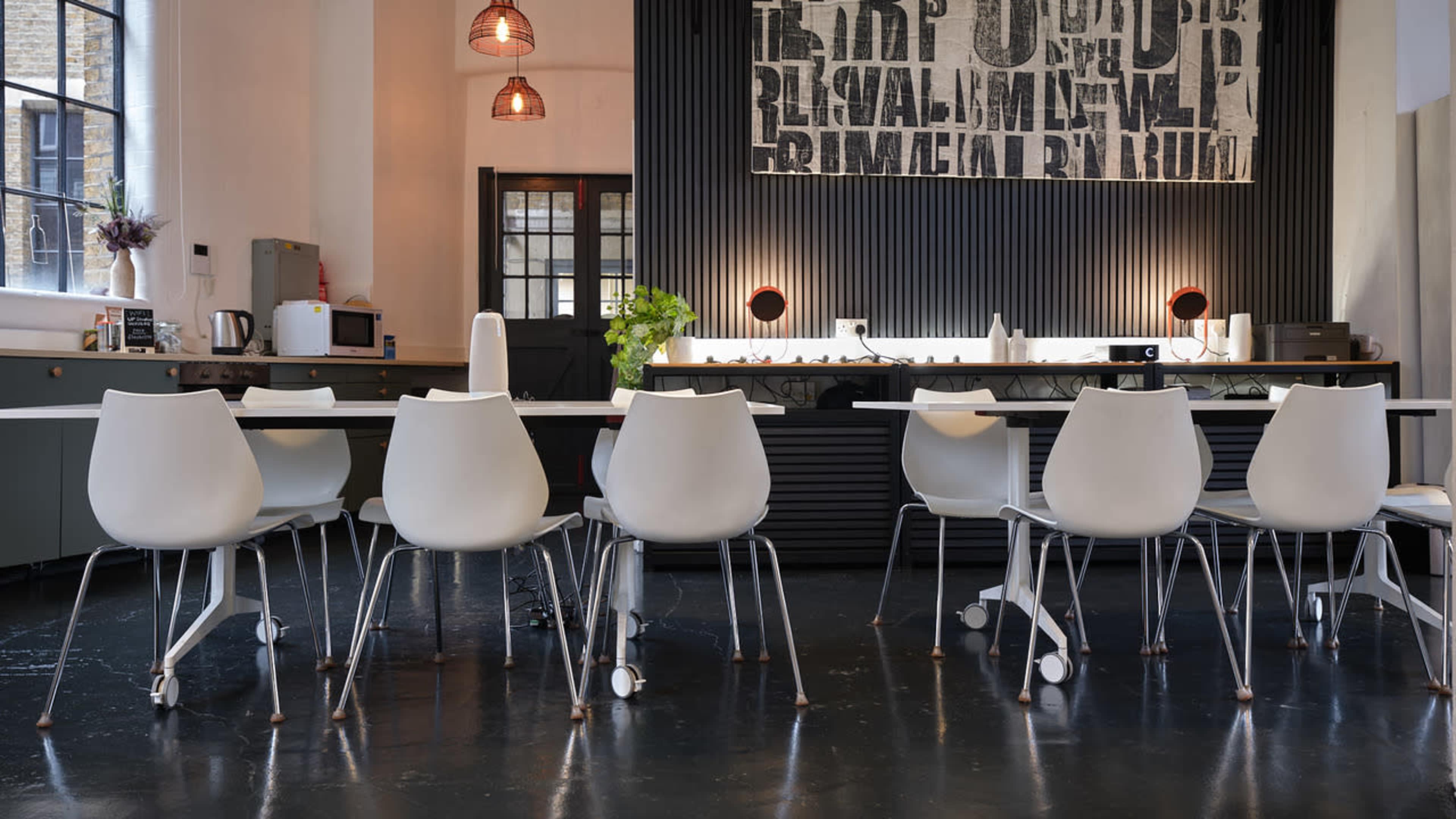 The image shows a modern kitchen space with a long table surrounded by white chairs, featuring a textured wall decor and pendant lights overhead.