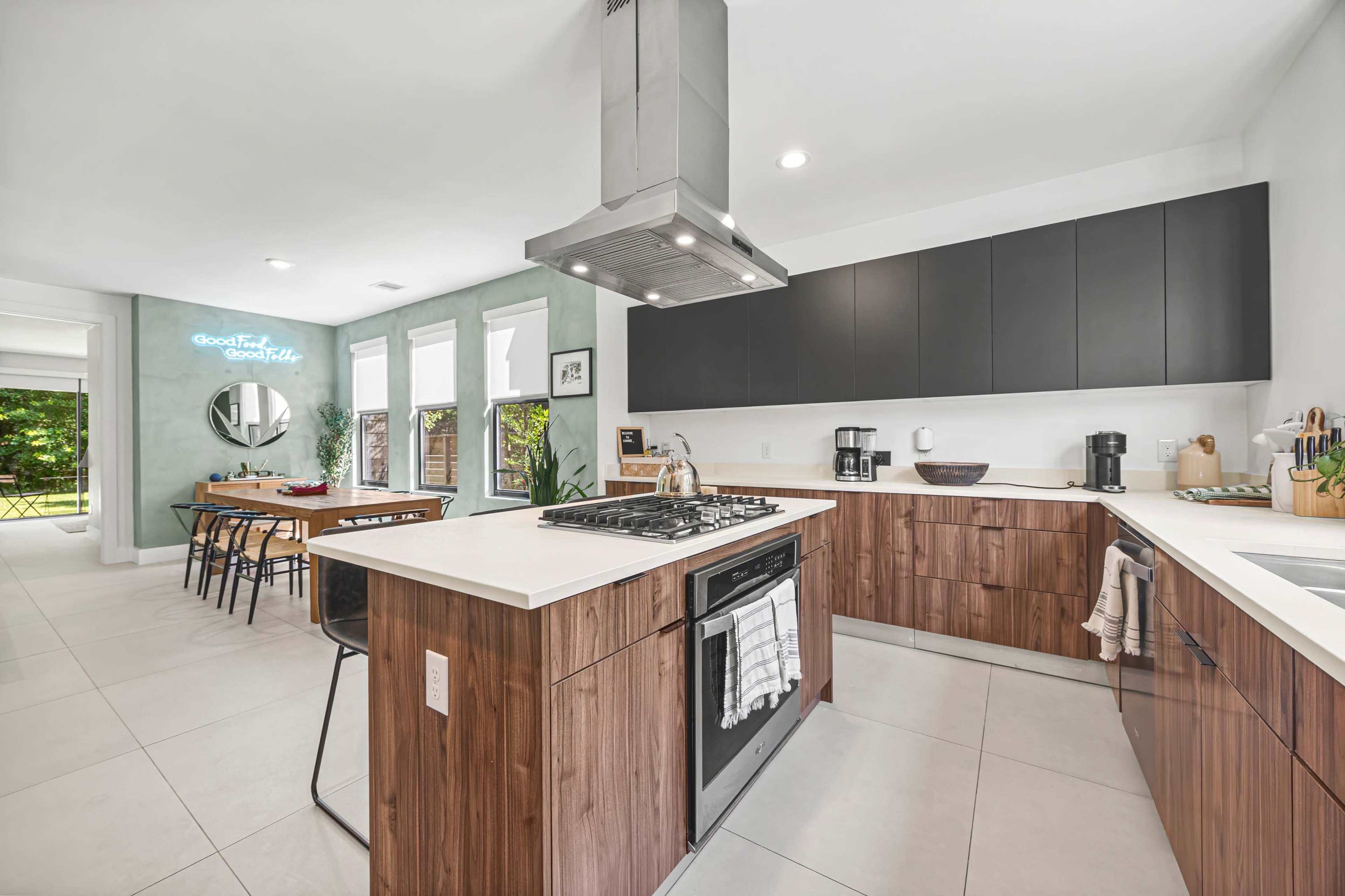 The image shows a modern kitchen featuring a central island with a gas stove, dark cabinetry, and an adjacent dining area with a wooden table and black chairs.
